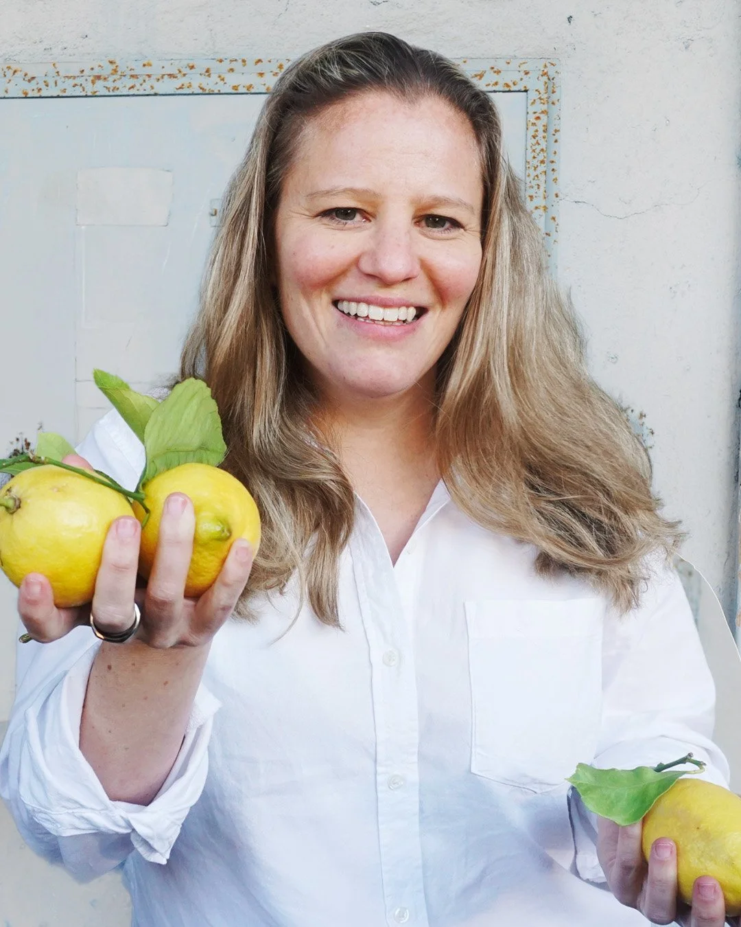 Woman with long blonde hair smiling and holding three yellow lemons with green leaves in front of a light-colored metal door.