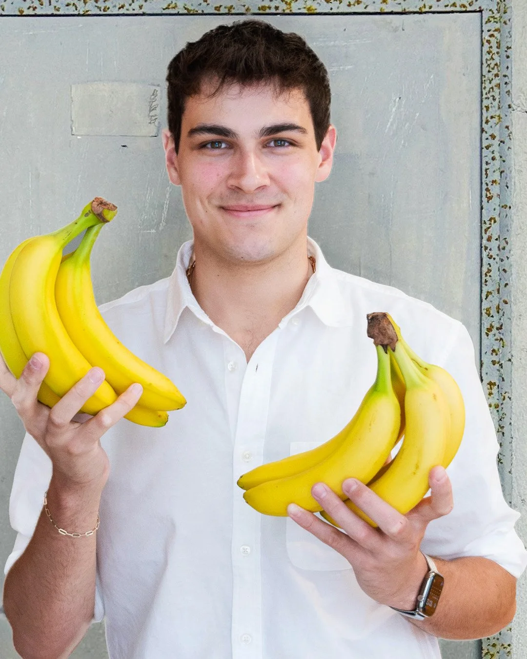 A young man with short dark hair, wearing a white shirt and a watch on his left wrist, holding two bananas in each hand, smiling at the camera.