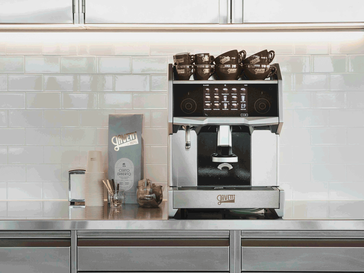 A modern espresso machine on a stainless steel counter with brown coffee cups on top, along with paper cups, a small container of stir sticks, a jar, and a menu board next to it, against a white tiled wall.