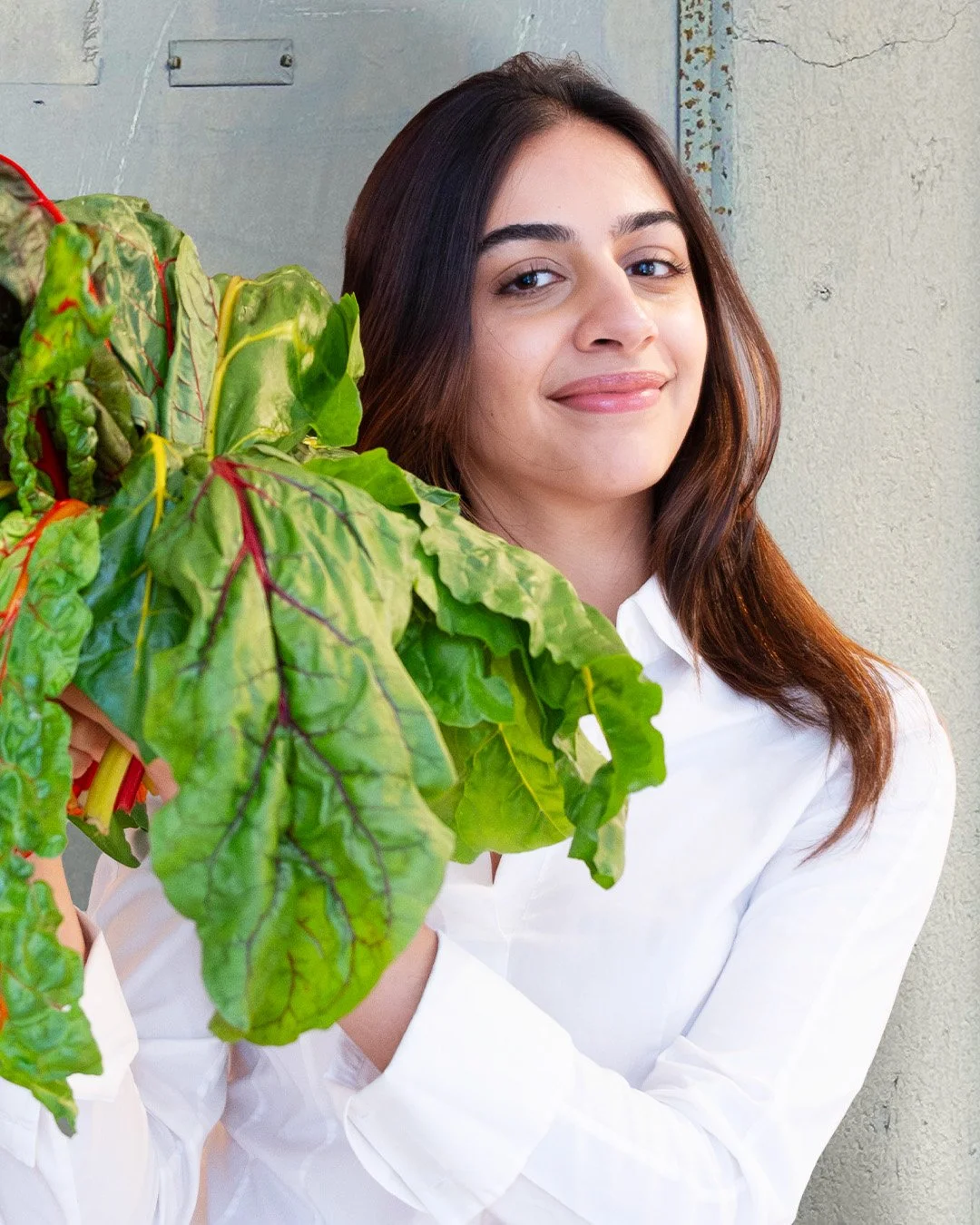 A woman with long brown hair smiling and holding a large Swiss chard plant with green and red-veined leaves, standing against a textured wall.