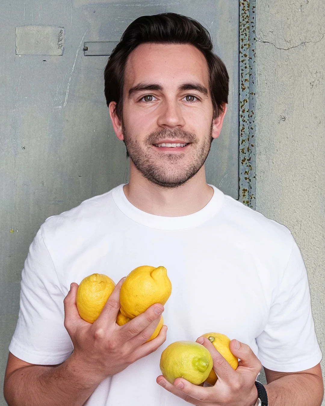 A man with dark hair and beard smiling is holding three yellow lemons in his right hand and two green lemons in his left hand. He is wearing a white t-shirt and is standing against a grayish textured wall.