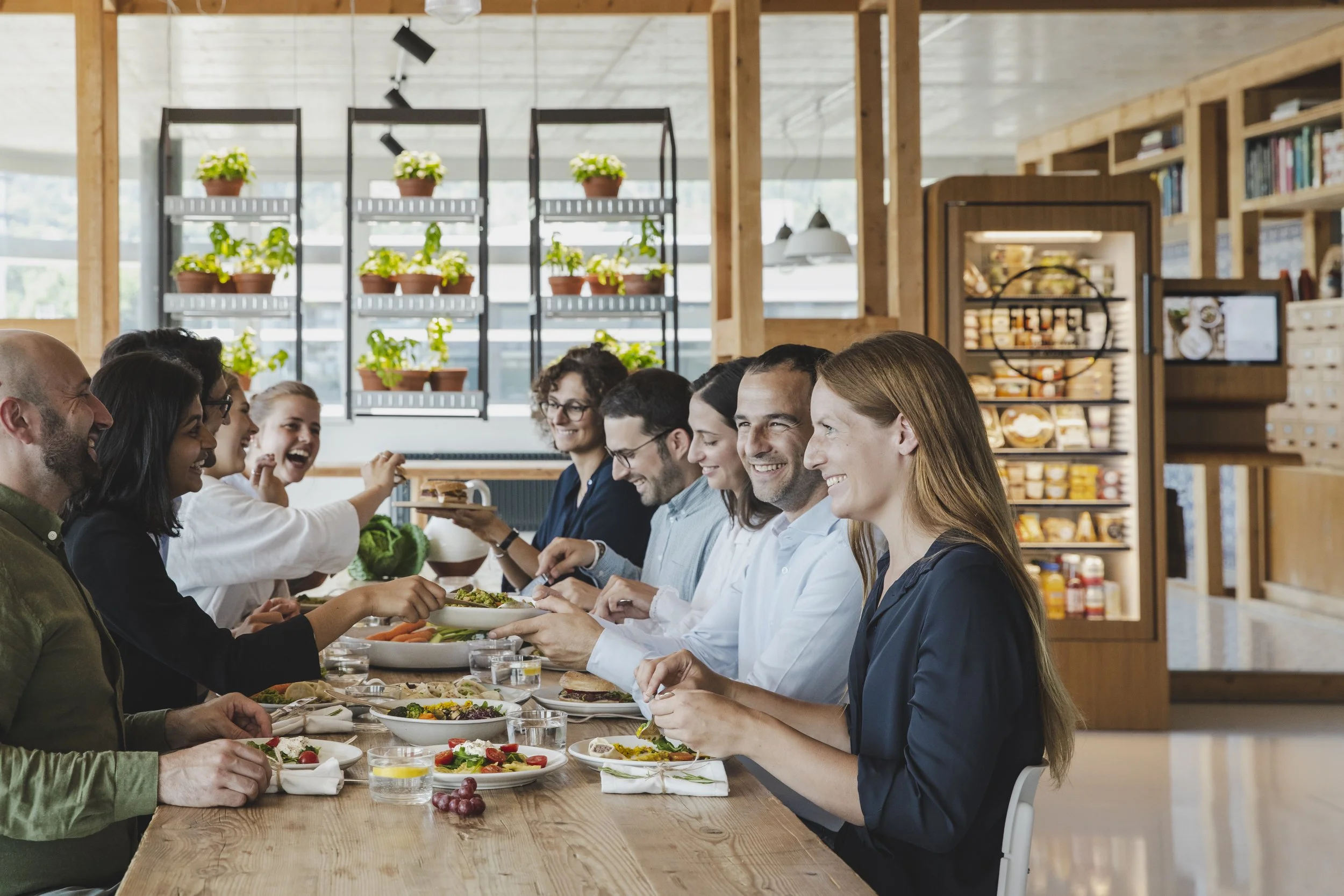 Group of people sitting at a long wooden table in a modern dining area, smiling and enjoying a meal together. The space has a bright, open atmosphere with potted plants on shelves in the background.