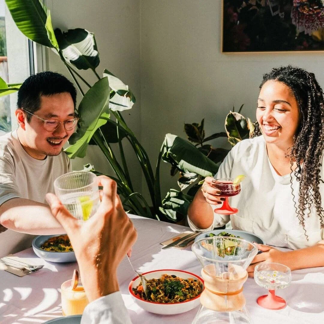 Three people enjoying a meal together at a table, with drinks and bowls of food, in a room with large green plants and sunlight.