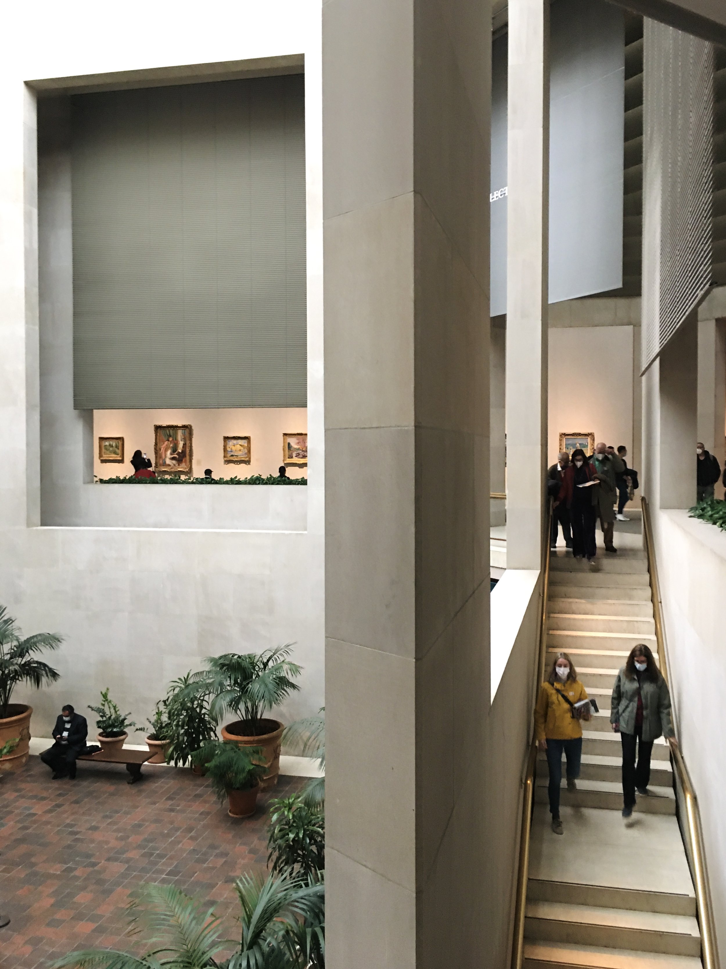 Interior view of a museum atrium with stairs, potted plants, and people walking and sitting, some wearing masks, and paintings displayed on the wall.
