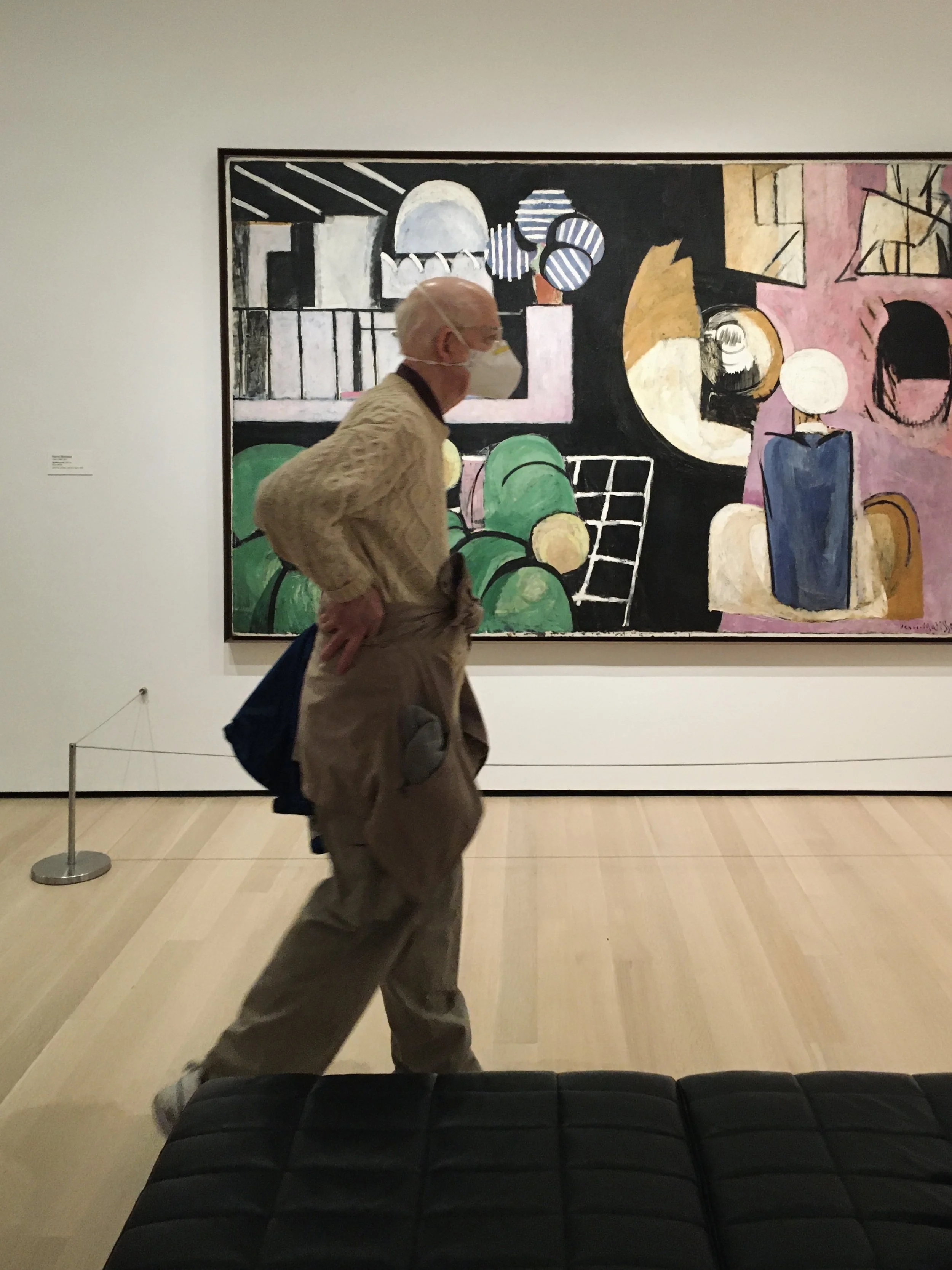 A person with white hair, wearing a beige cable-knit sweater, khaki pants, a face mask, and carrying a blue bag, walking past a colorful abstract painting in an art gallery.