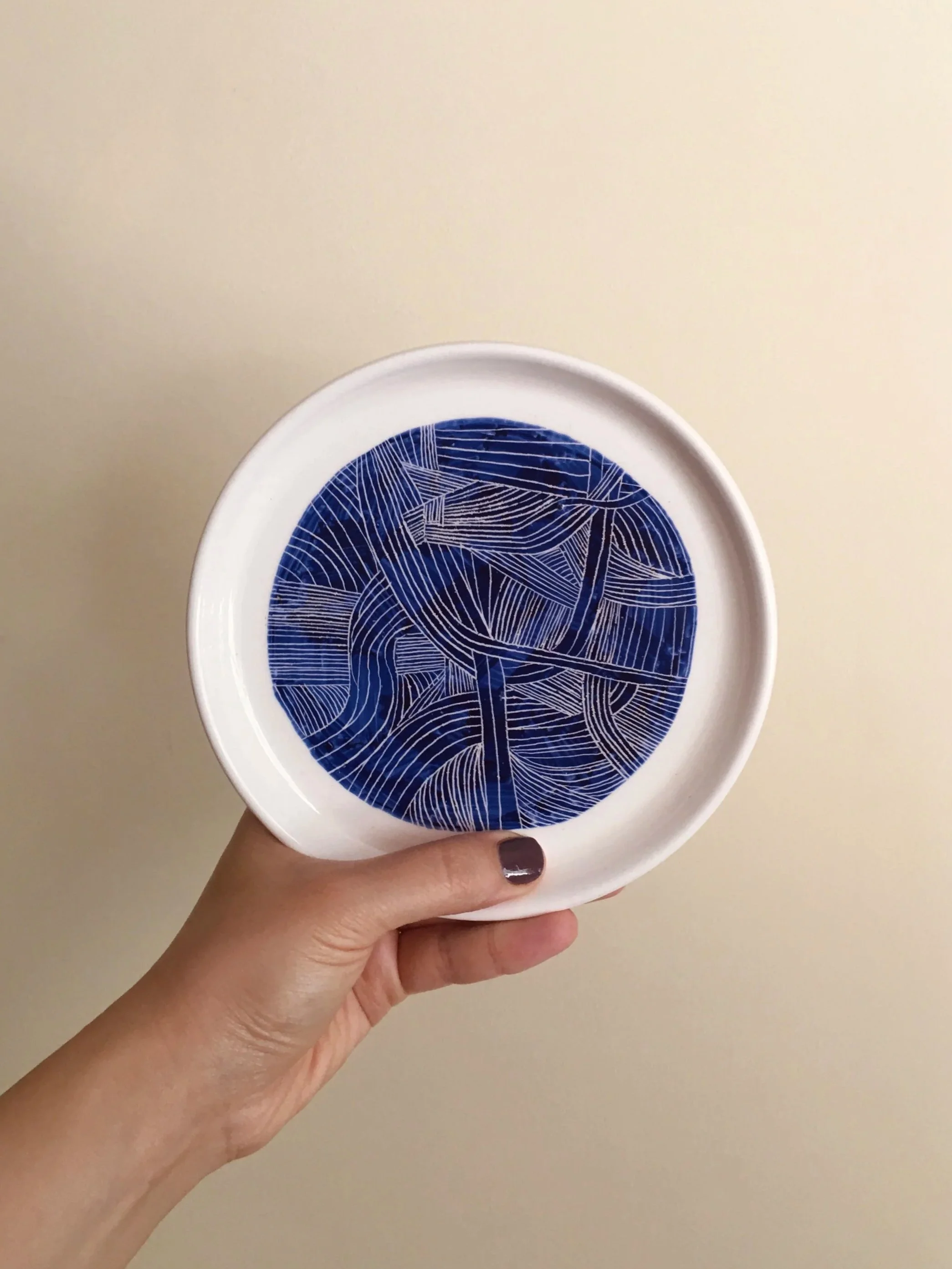 A hand holding a white ceramic plate with a blue geometric pattern design on the inside, against a plain beige background.