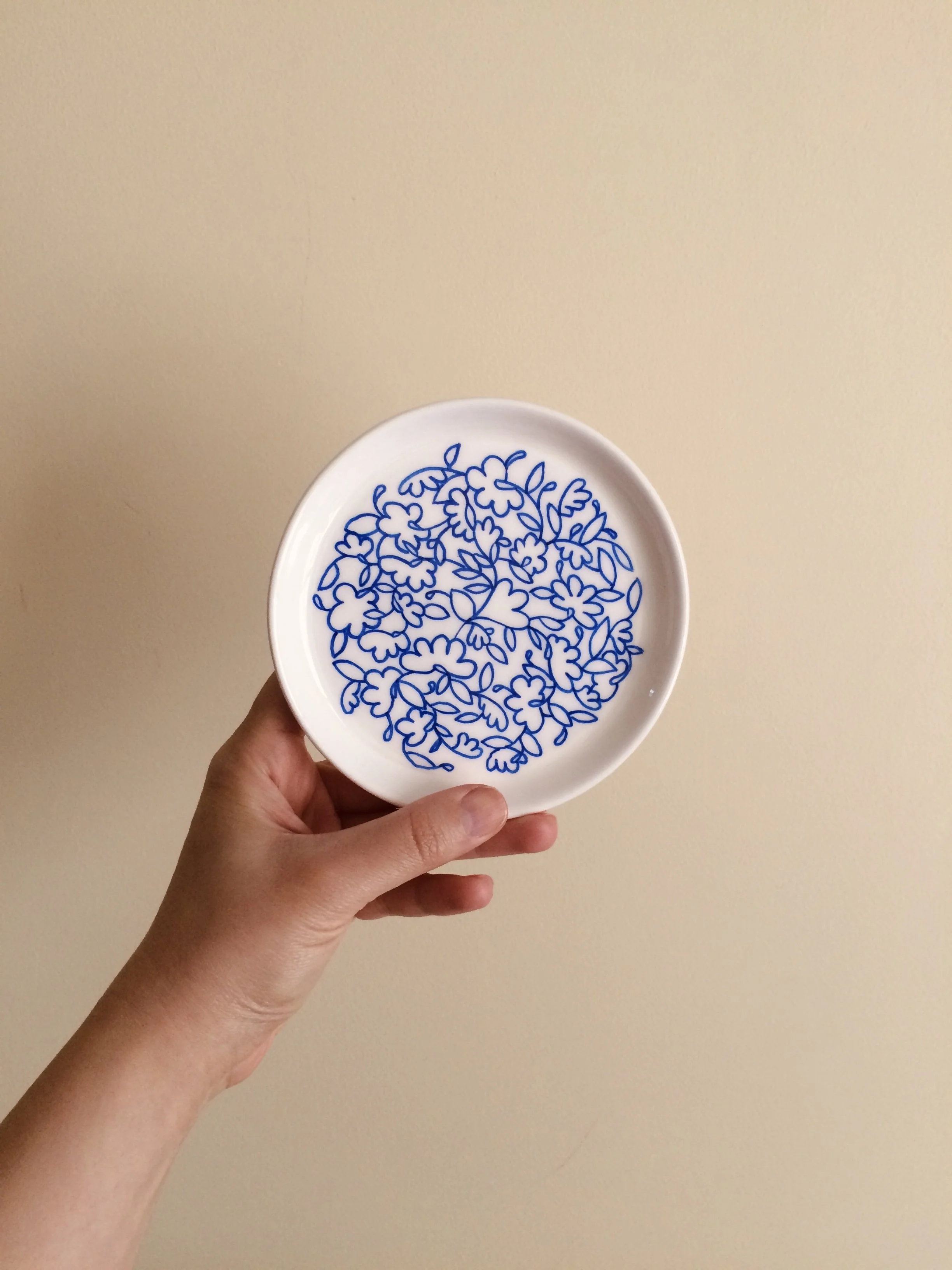 A hand holding a white plate with a blue floral pattern on a plain beige background.