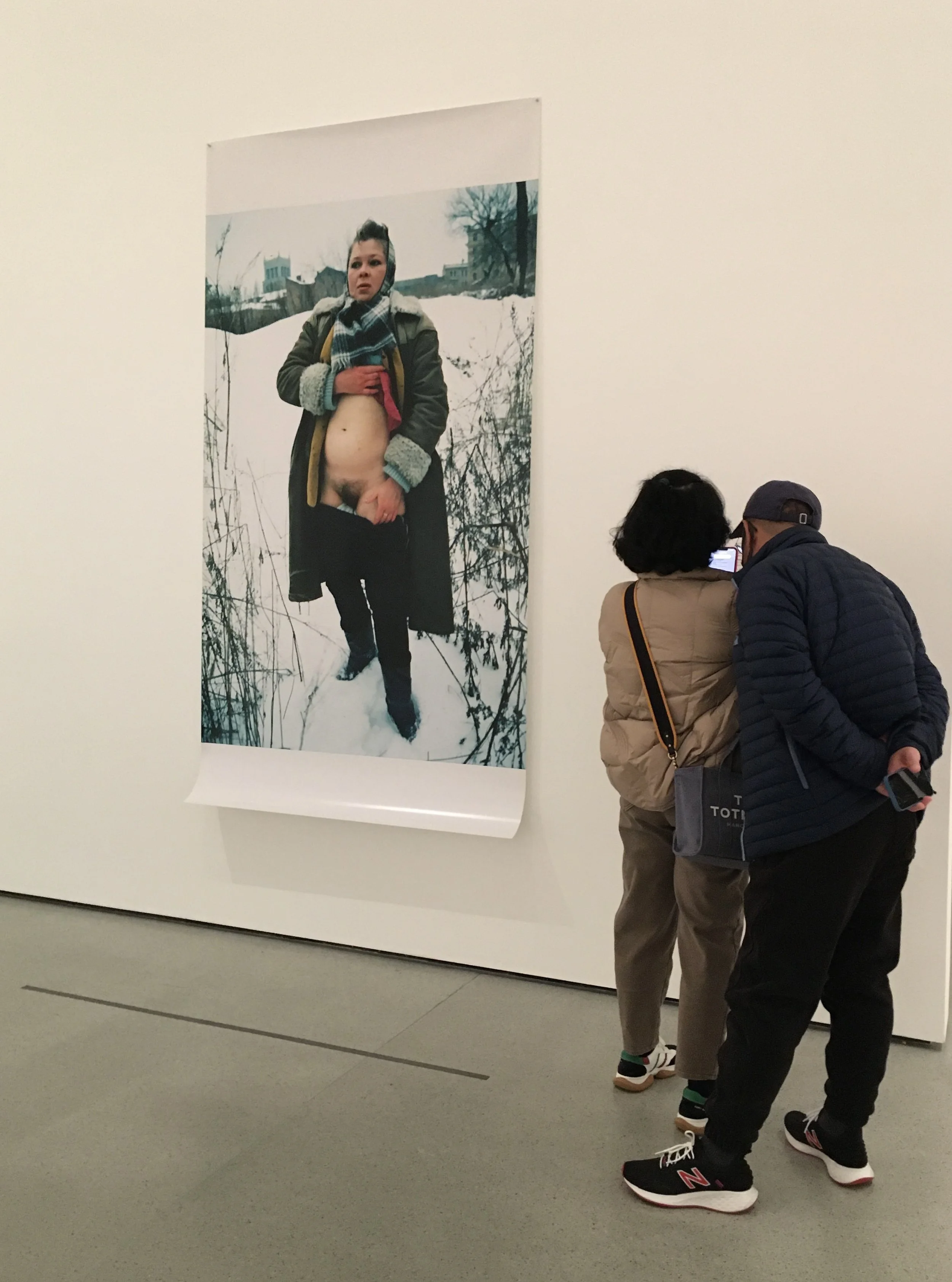 Two people are looking at a large art photograph displayed in a gallery. The photograph features a woman standing outdoors in snow, wearing a coat with the front open, revealing her bare chest and stomach, and holding her shirt with one hand.