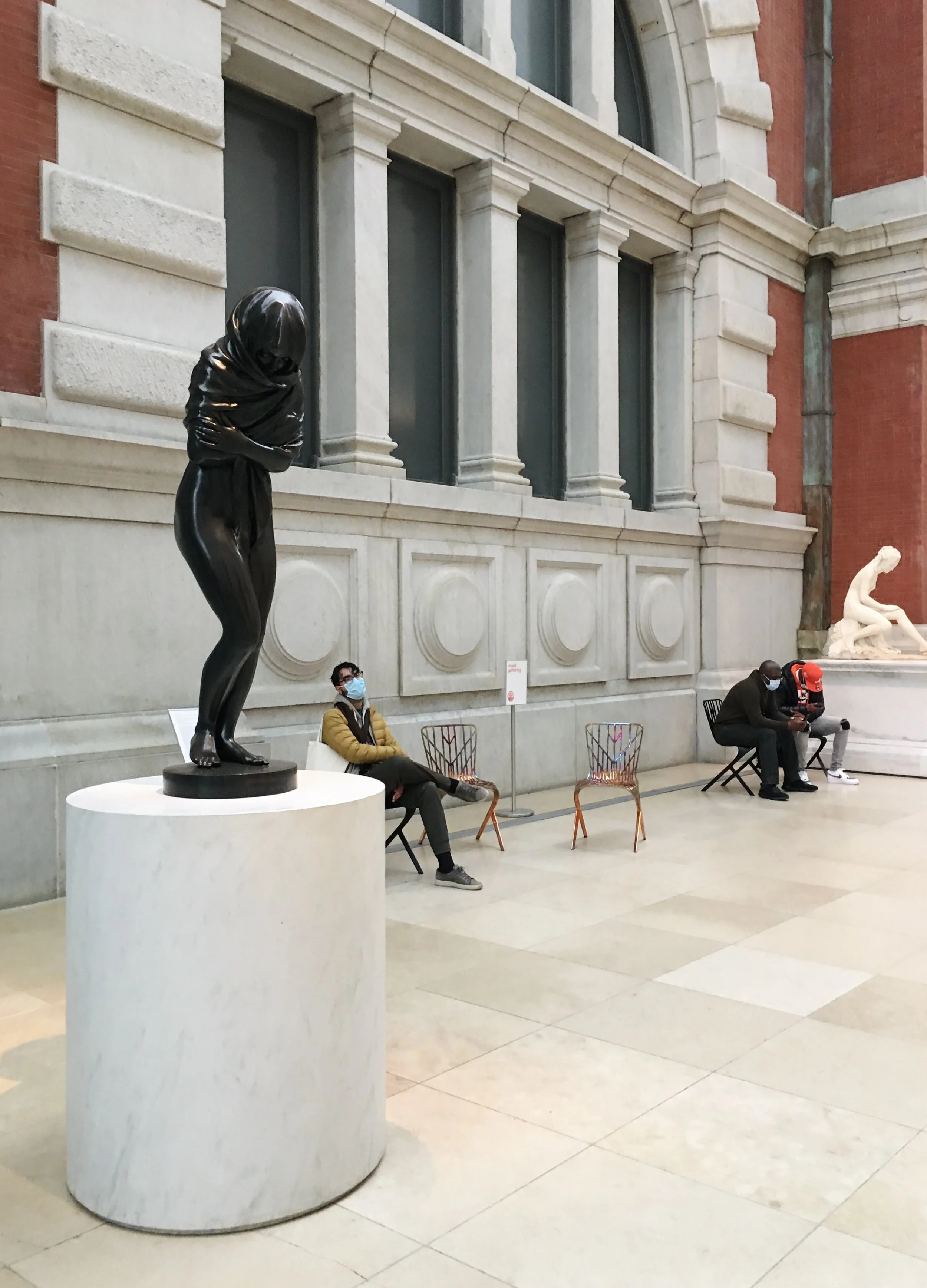 Inside a museum, a black sculpture of a frightened child stands on a white pedestal to the left, while three people sit on chairs apart, all wearing face masks.