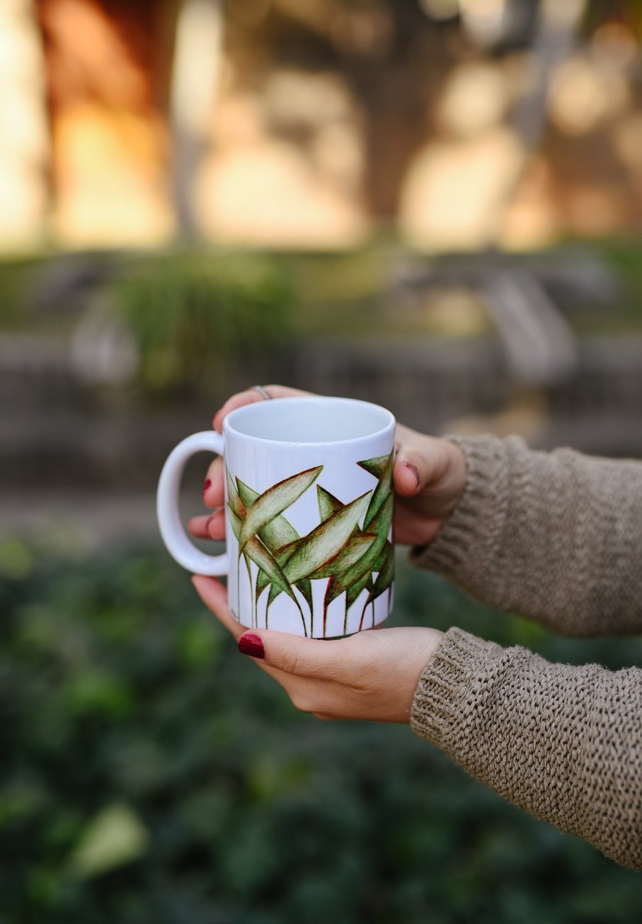 Person holding a white ceramic mug with a green leafy plant design, outdoors in a blurred garden or park background.