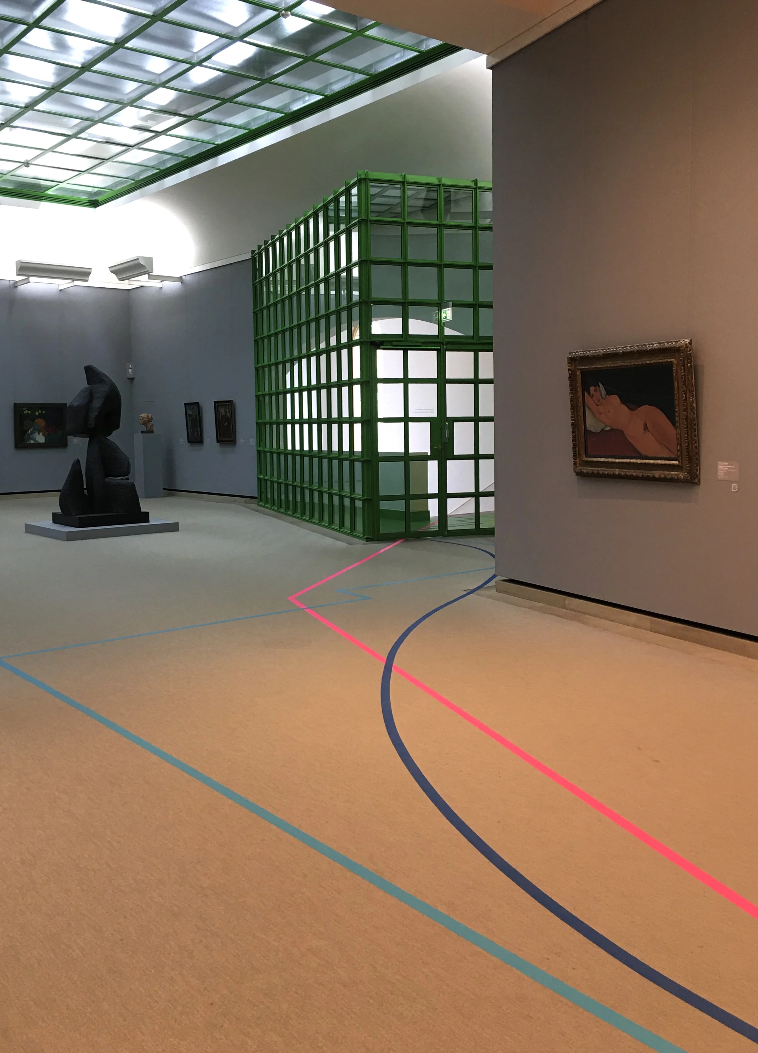 Interior view of an art gallery or museum with sculptures, paintings, and a green lattice structure. The ceiling has a glass or plastic roof, and the floor has colorful lines.