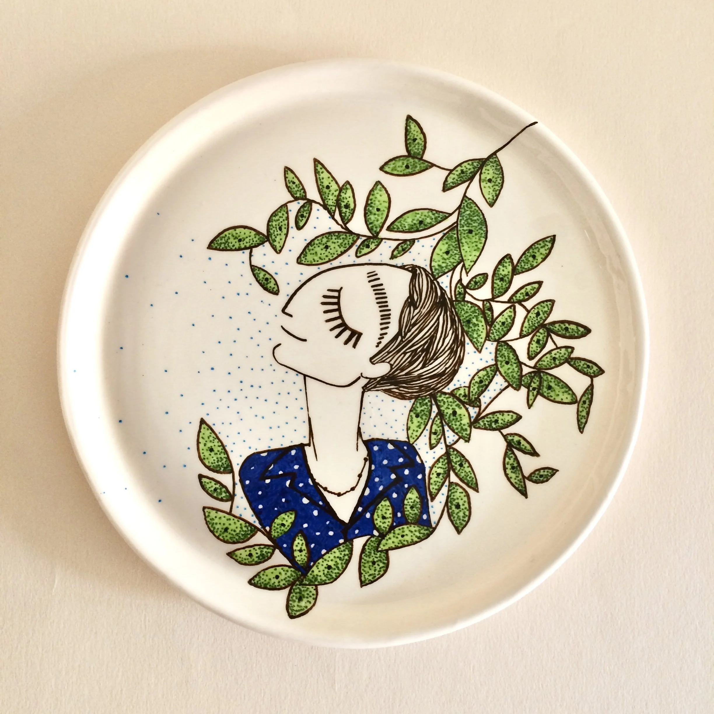 Decorative round ceramic plate with a line art illustration of a woman with closed eyes, short hair, and a blue polka-dot top, surrounded by green leafy branches and small blue dots.