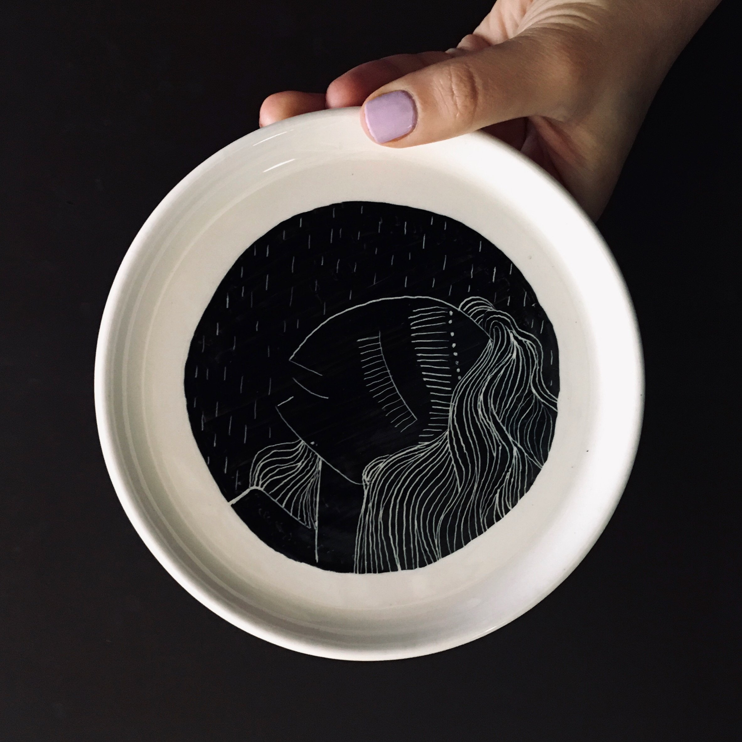 A top-down view of a white bowl with black interior featuring a white abstract line drawing of a face and flowing hair on a dark surface, held by a hand with light purple painted nails.