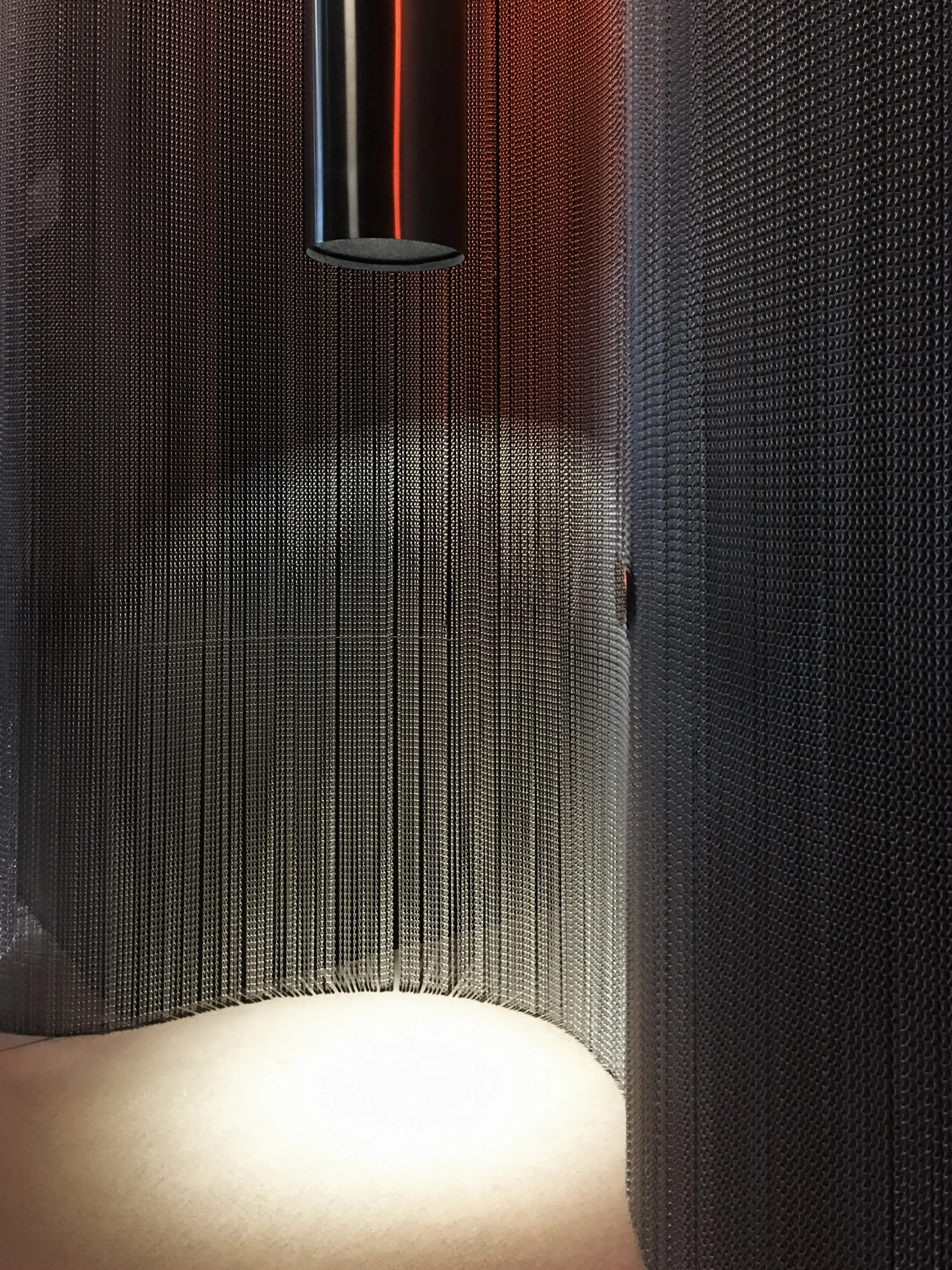 Part of a modern elevator interior with hanging black metal mesh panels, a round black ceiling light, and red lighting accents.