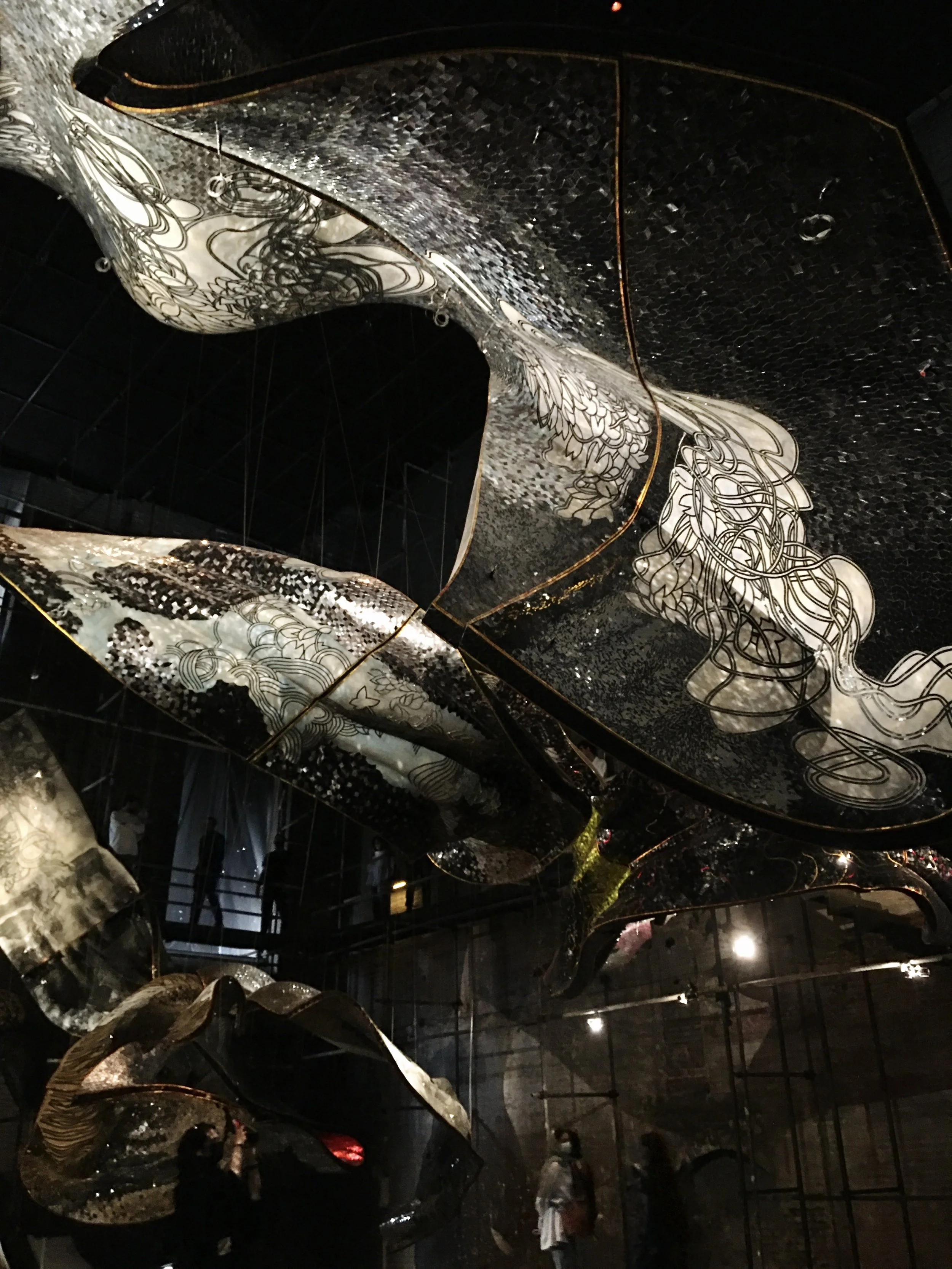A large, abstract art installation with flowing, layered, reflective surfaces and intricate line drawings, hanging from the ceiling in a dark room.