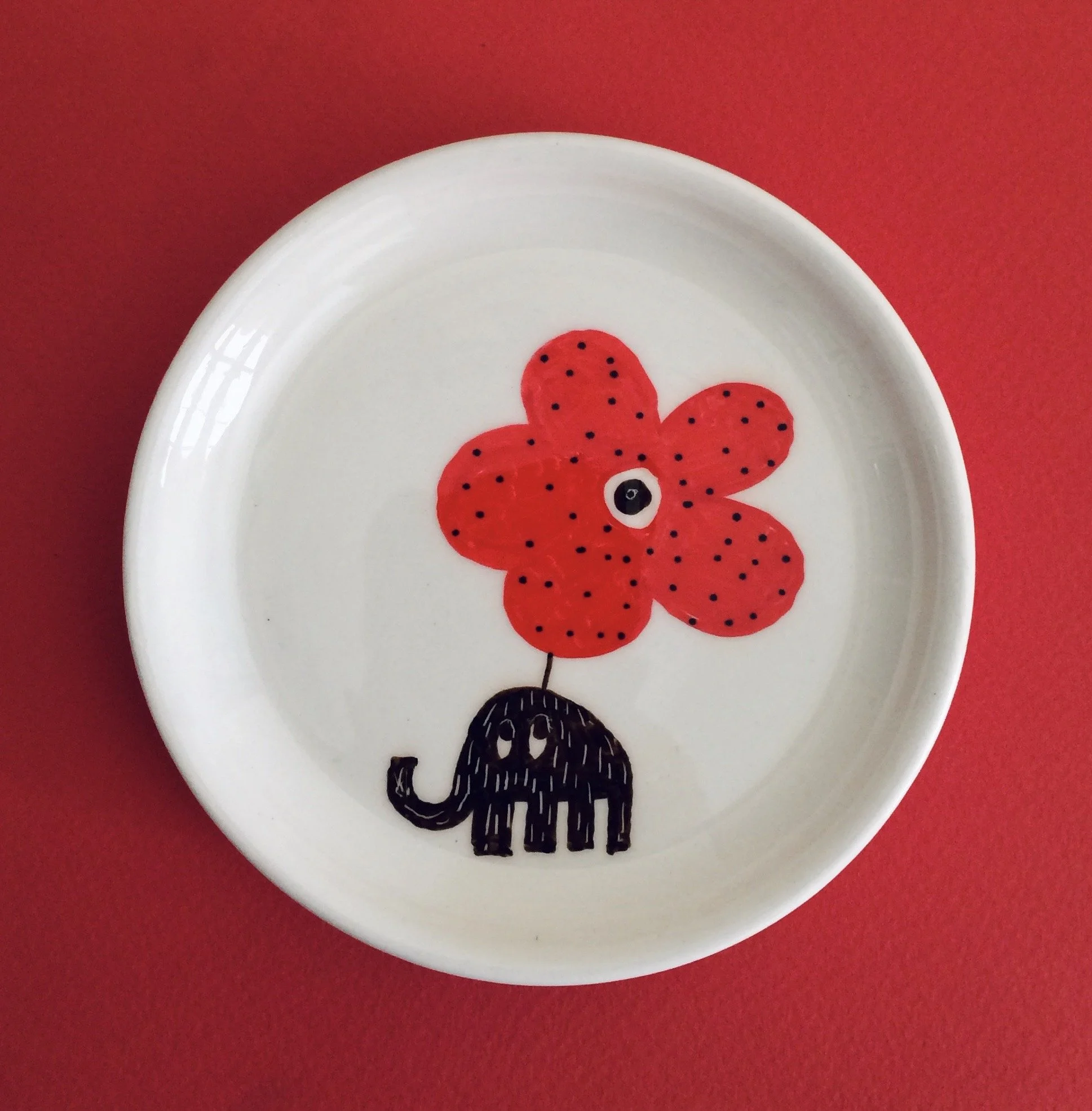 A white ceramic plate with a painted red flower with black dots, featuring a cartoonish eye, connected to a small black elephant with white eyes and a tail, set against a red background.