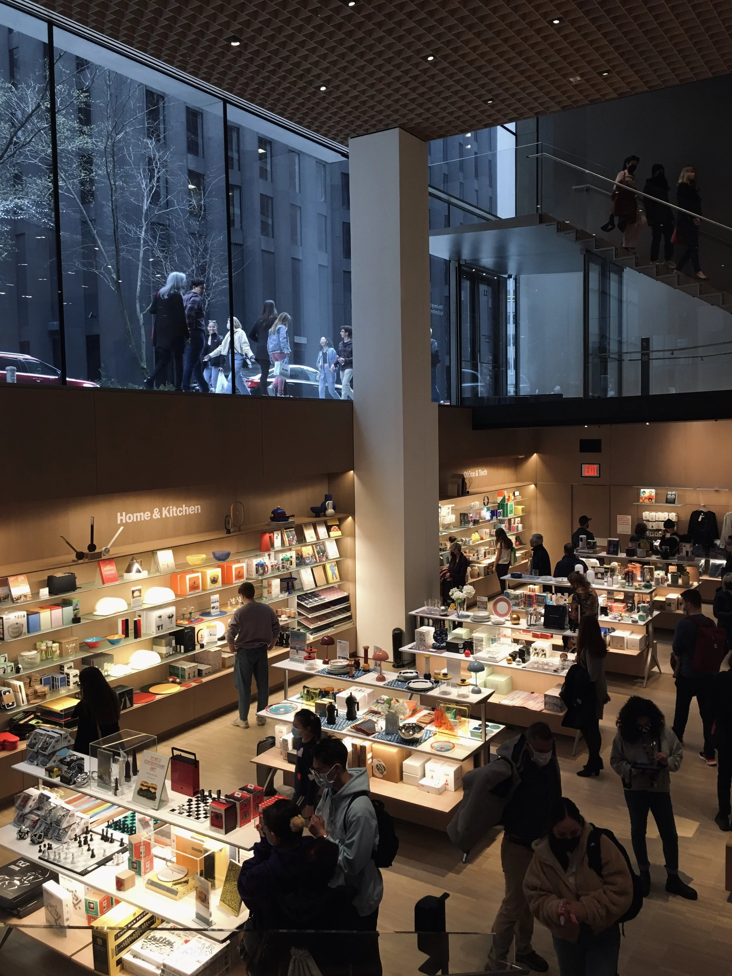 Interior of a retail store with a section labeled 'Home & Kitchen' displaying various household items, shoppers browsing, and a large glass window showing people outside.