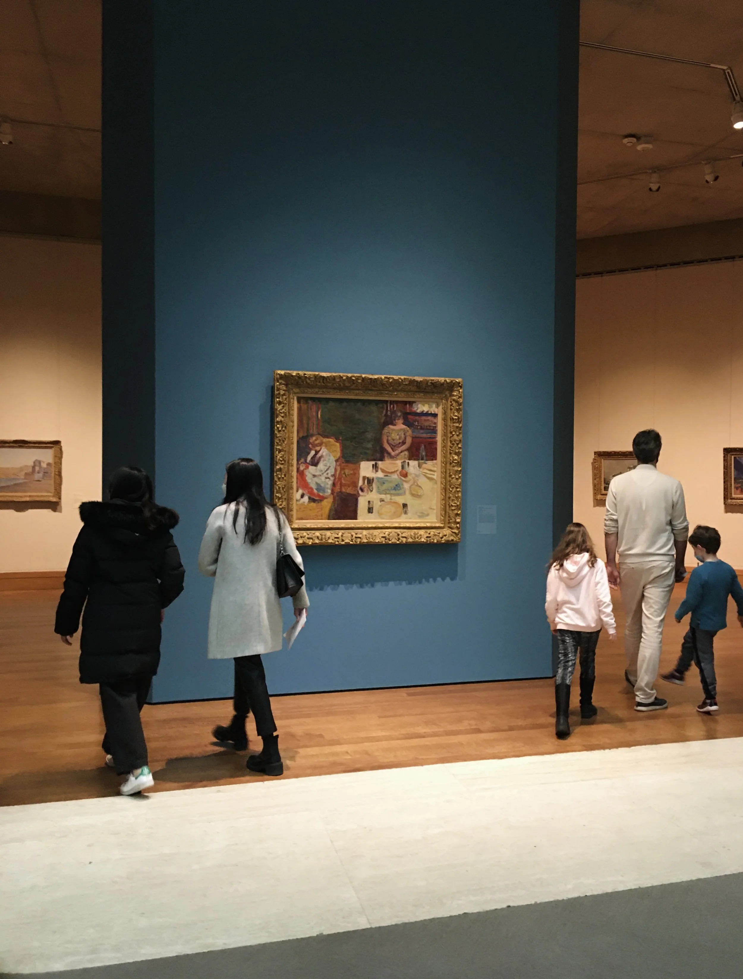 People walking in an art museum, viewing paintings on display. The central painting is a colorful, framed artwork on a blue wall.
