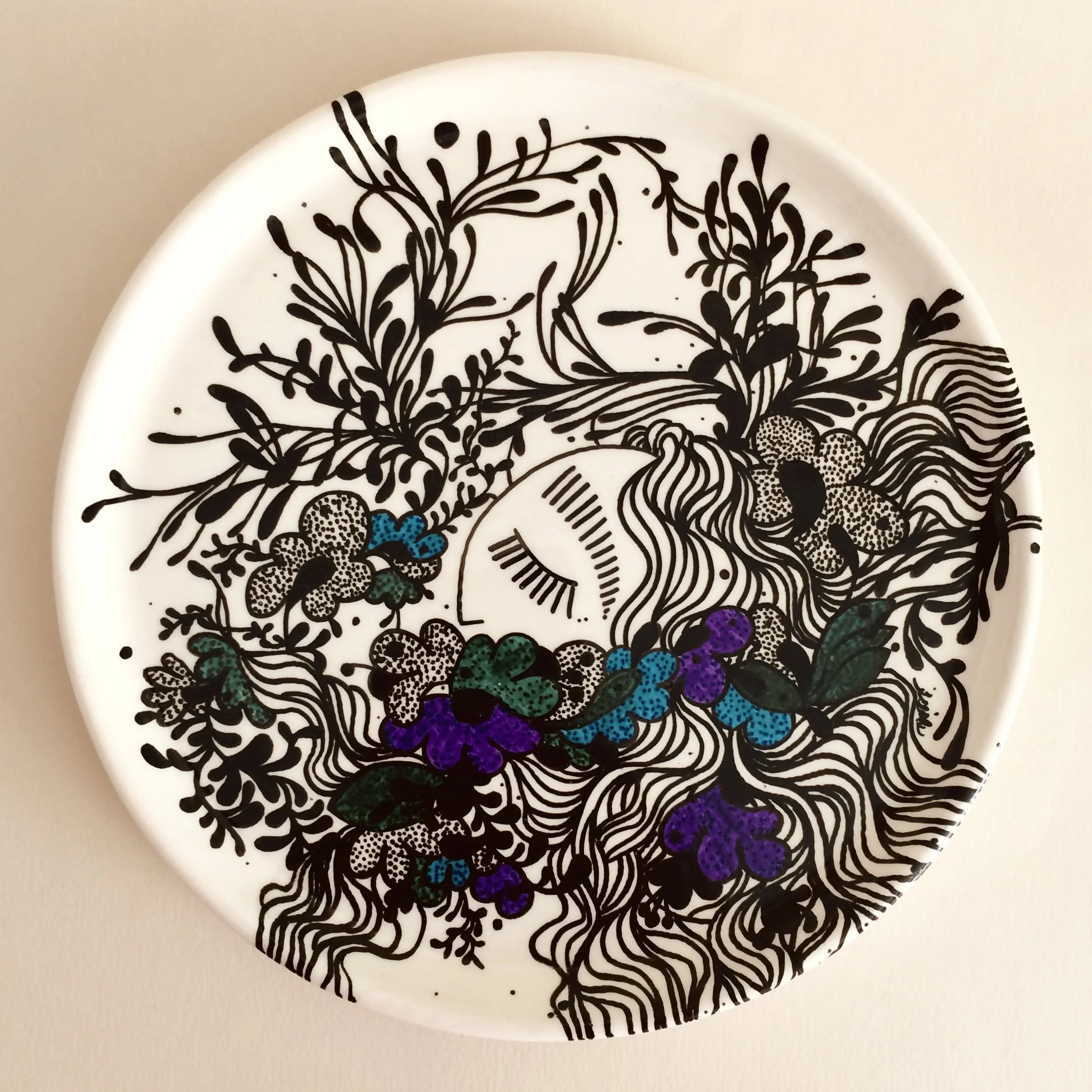 Decorative plate with a black and white illustration of a woman's face surrounded by flowers, leaves, and flowing hair, with some colored accents in purple, teal, and green.