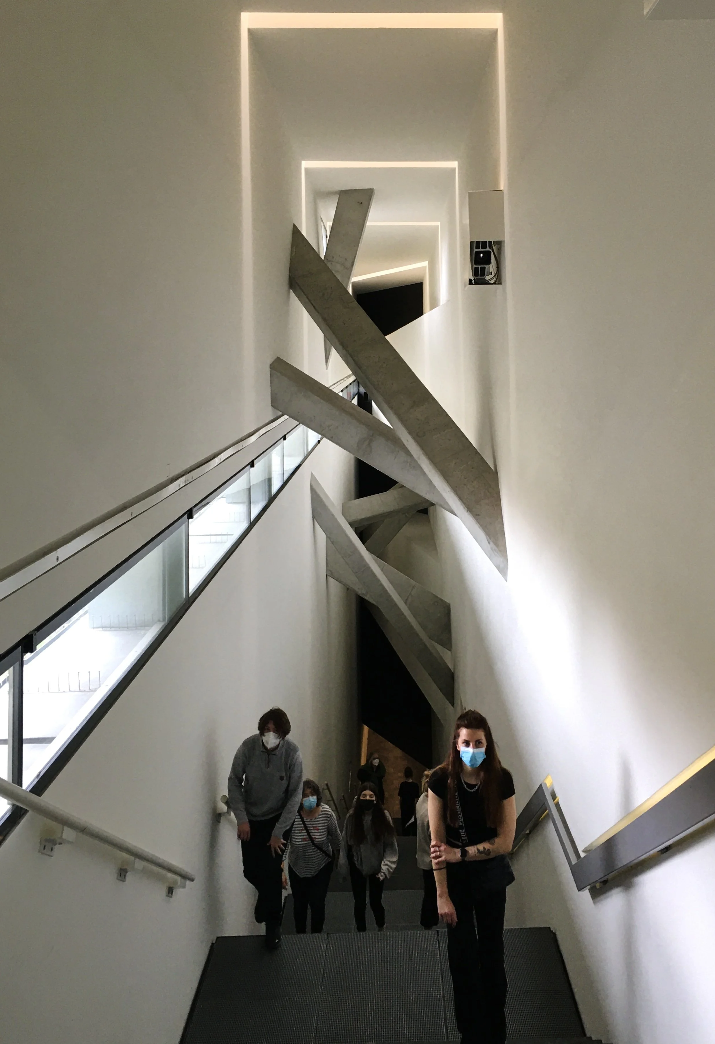 People wearing masks walking up a staircase inside a modern building with unique concrete architectural features and illuminated ceiling design.