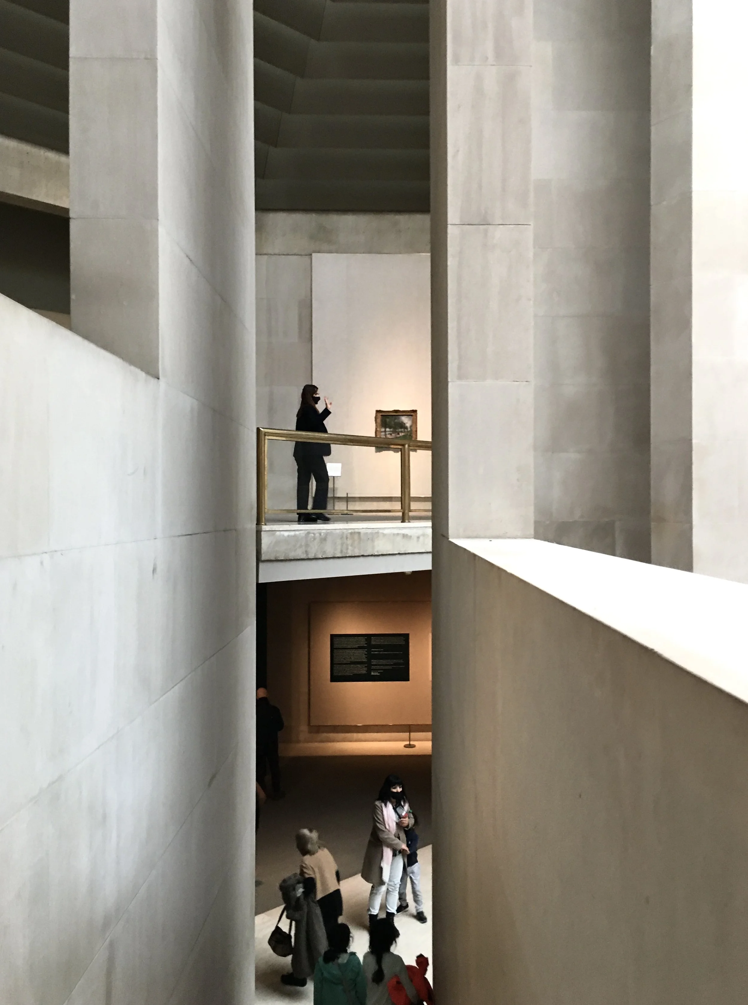 People inside a modern art museum, with one person on an upper level taking a photo and a group of visitors on the lower level near a wall with an informational plaque.