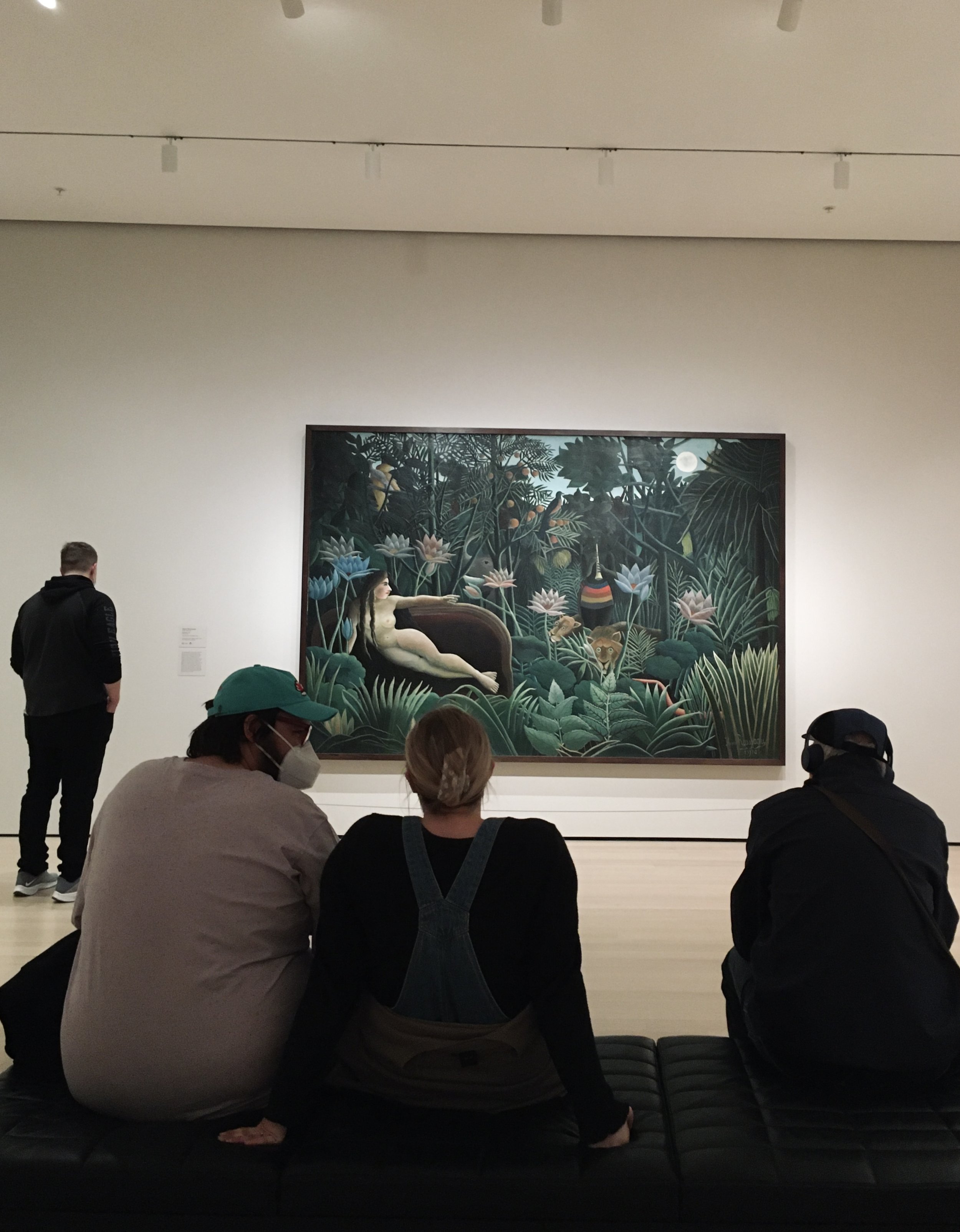 People sitting on a bench in front of a large painting of a jungle scene with a reclining woman, plants, animals, and a moon.