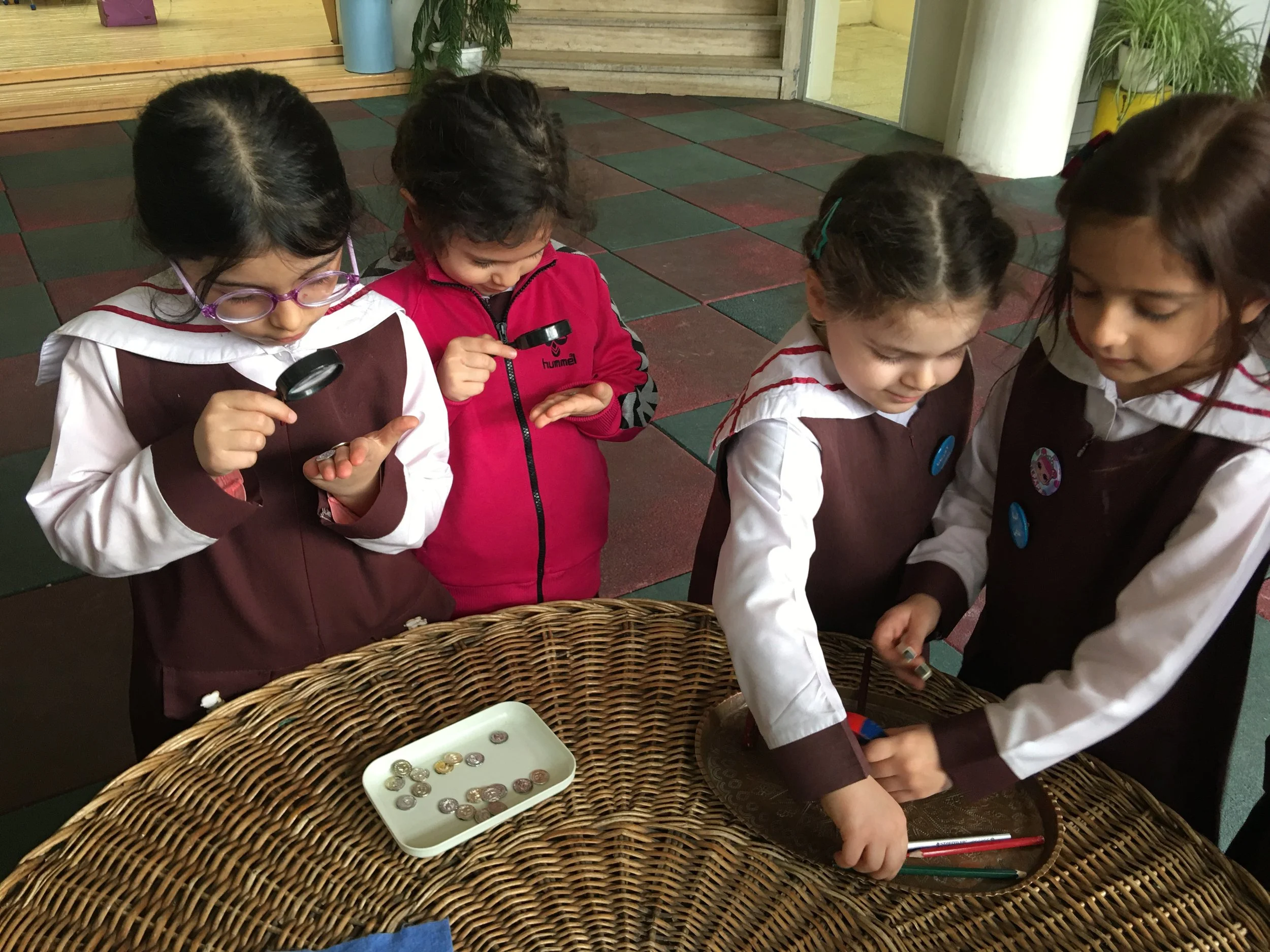 Four young girls, dressed in school uniforms, are gathered around a wicker table, examining coins and other small objects with magnifying glasses.