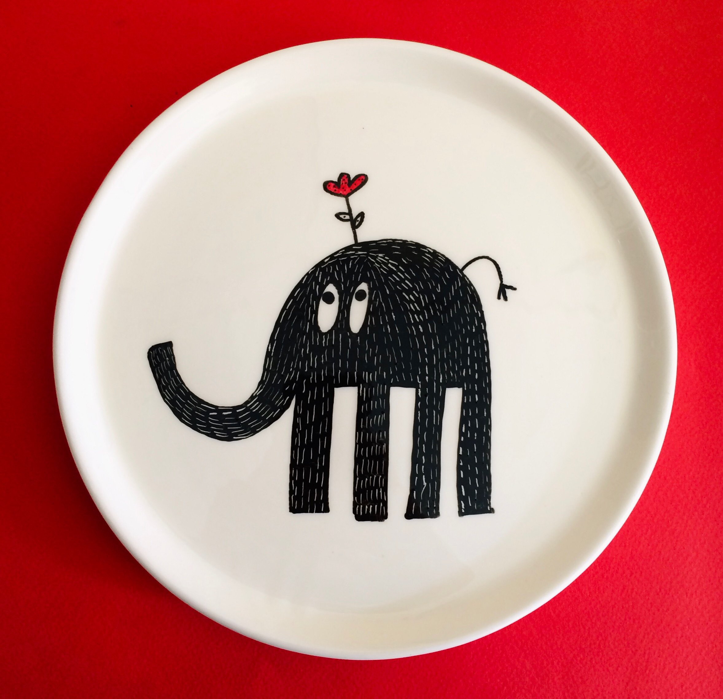 Decorative white plate with a black cartoon elephant illustration with a small red heart-shaped flower on its head, standing on a red surface.