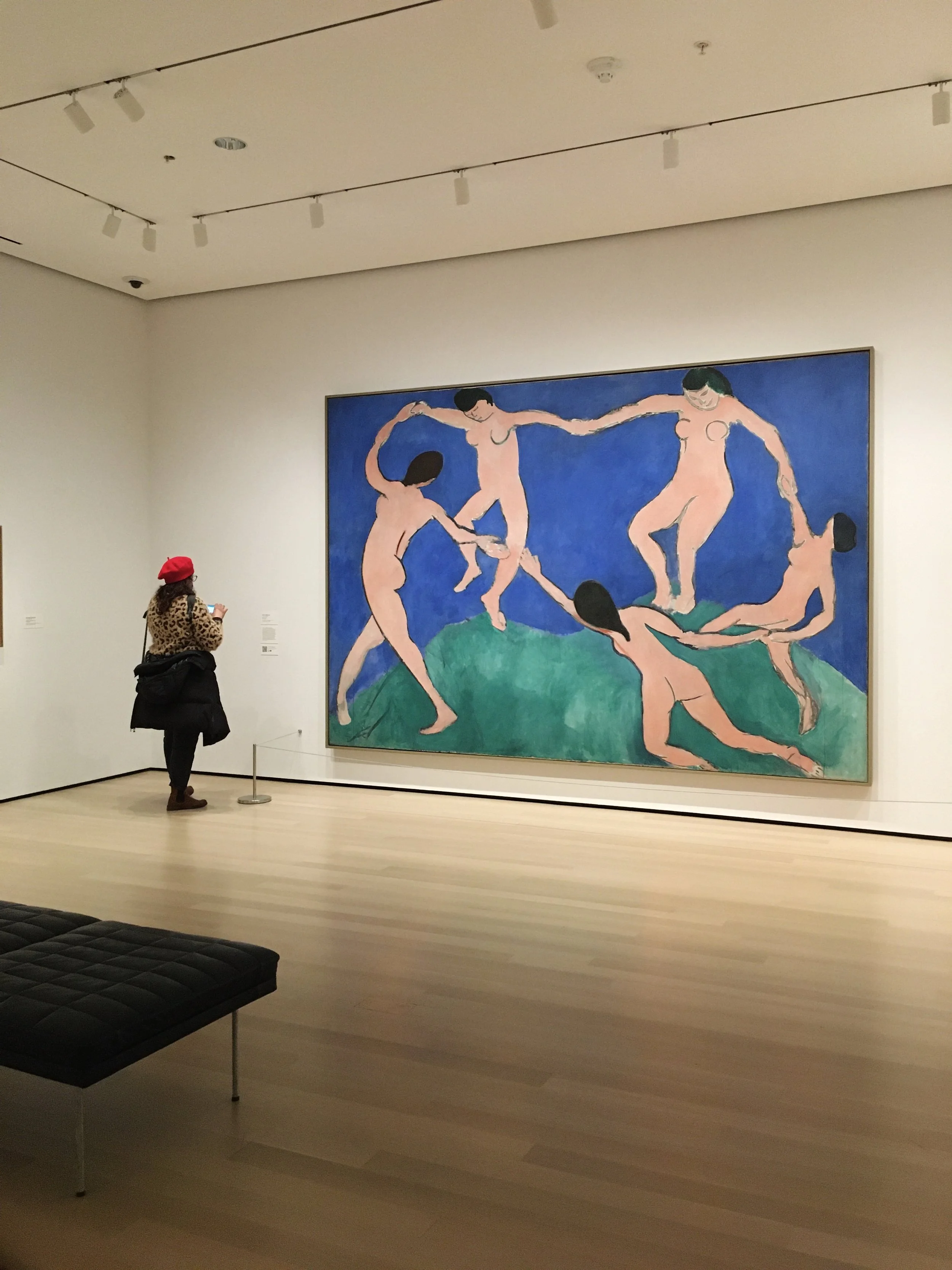 A person wearing a leopard print jacket and a red hat is looking at a large painting in an art gallery. The painting features abstracted nude figures with black hair and exaggerated proportions, in a dance-like formation on a blue background.