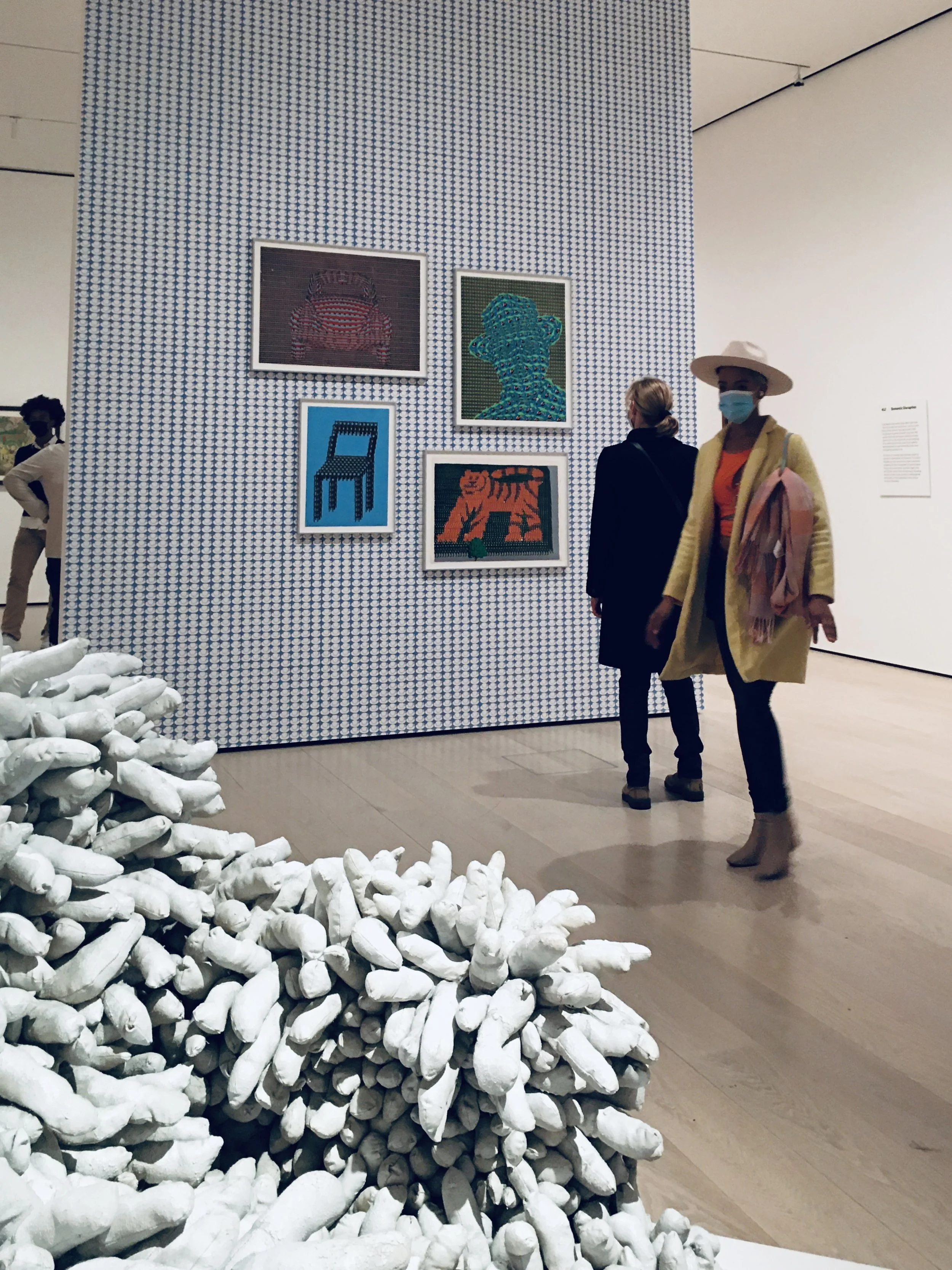 People viewing modern art in a gallery with a textured blue and white wall, featuring framed pixelated style artworks of animals and a chair; white textile sculpture in the foreground; woman in yellow coat and wide-brim hat wearing face mask.