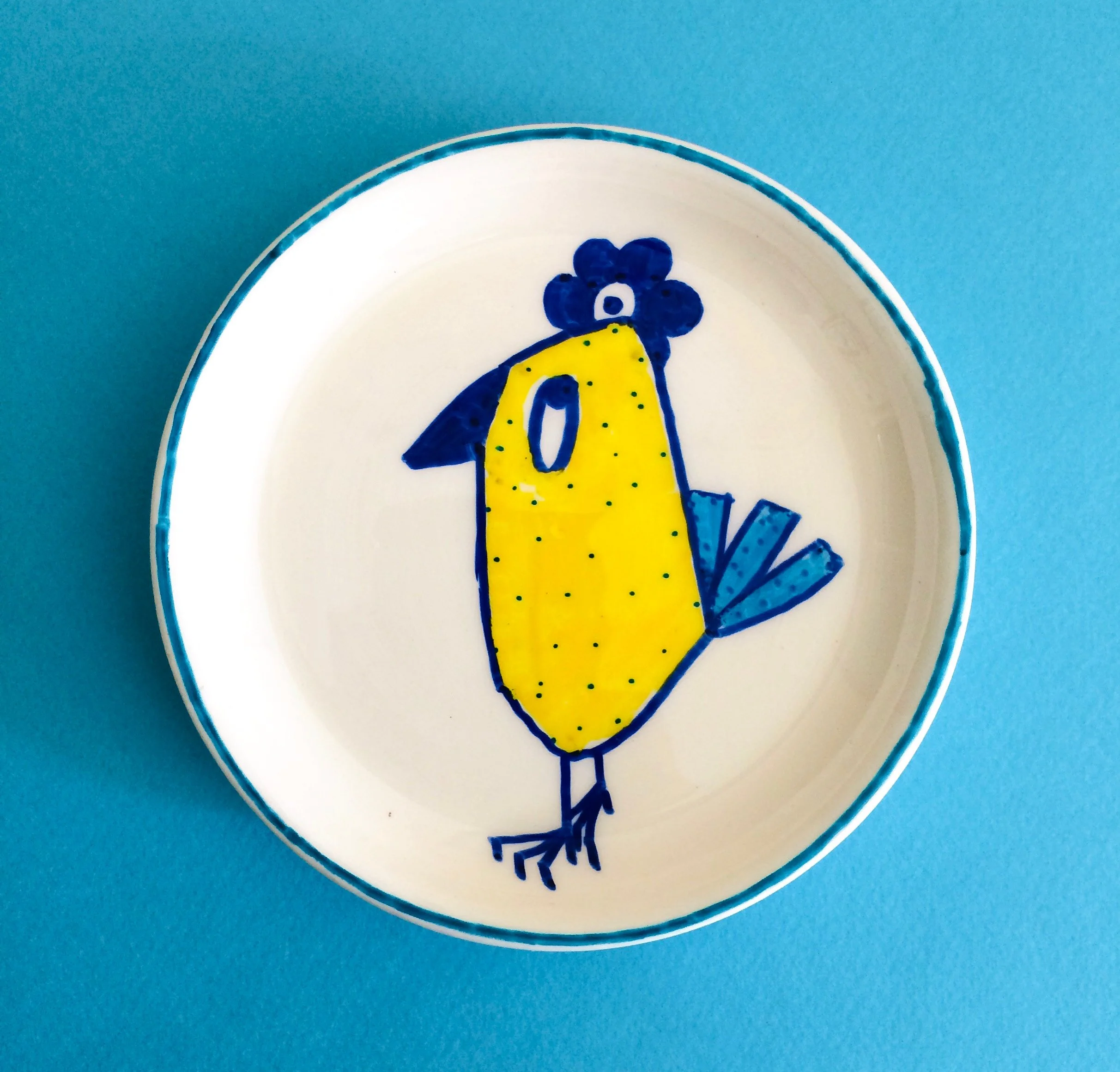 Decorative plate featuring a colorful, cartoon-style bird illustration, with a yellow body, blue wings, tail, and head, on a blue background.