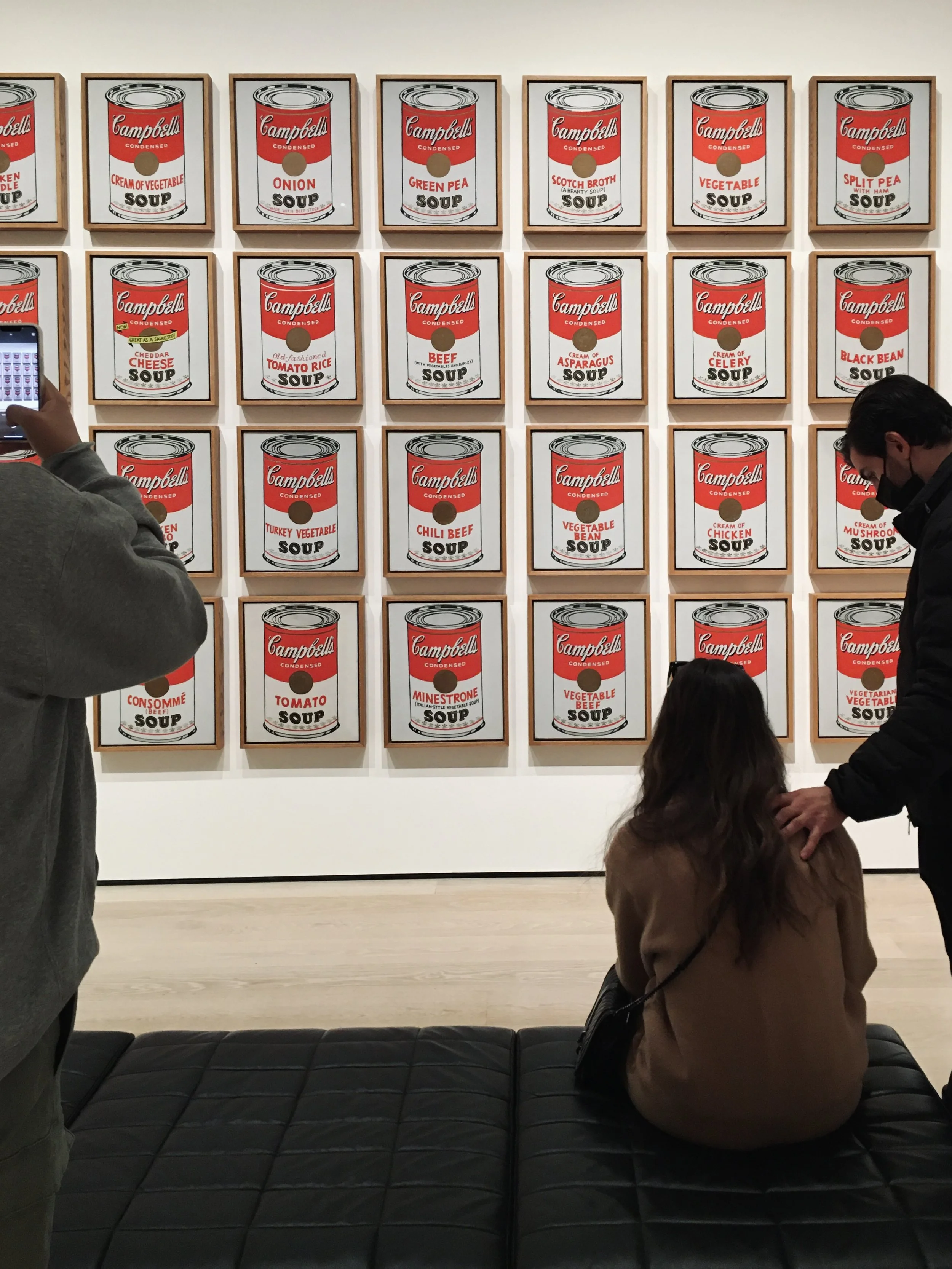 Gallery featuring framed art of Campbell's soup cans, with visitors observing and taking photos.