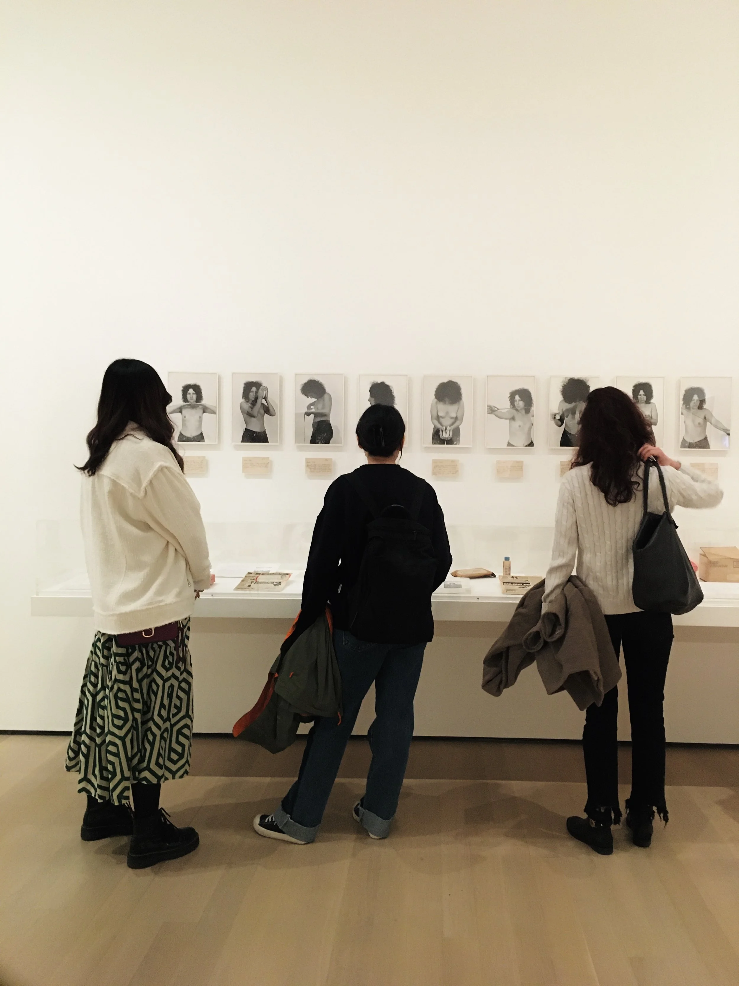 Three women viewing an art exhibit of black-and-white photographs of a woman with curly hair at a gallery.