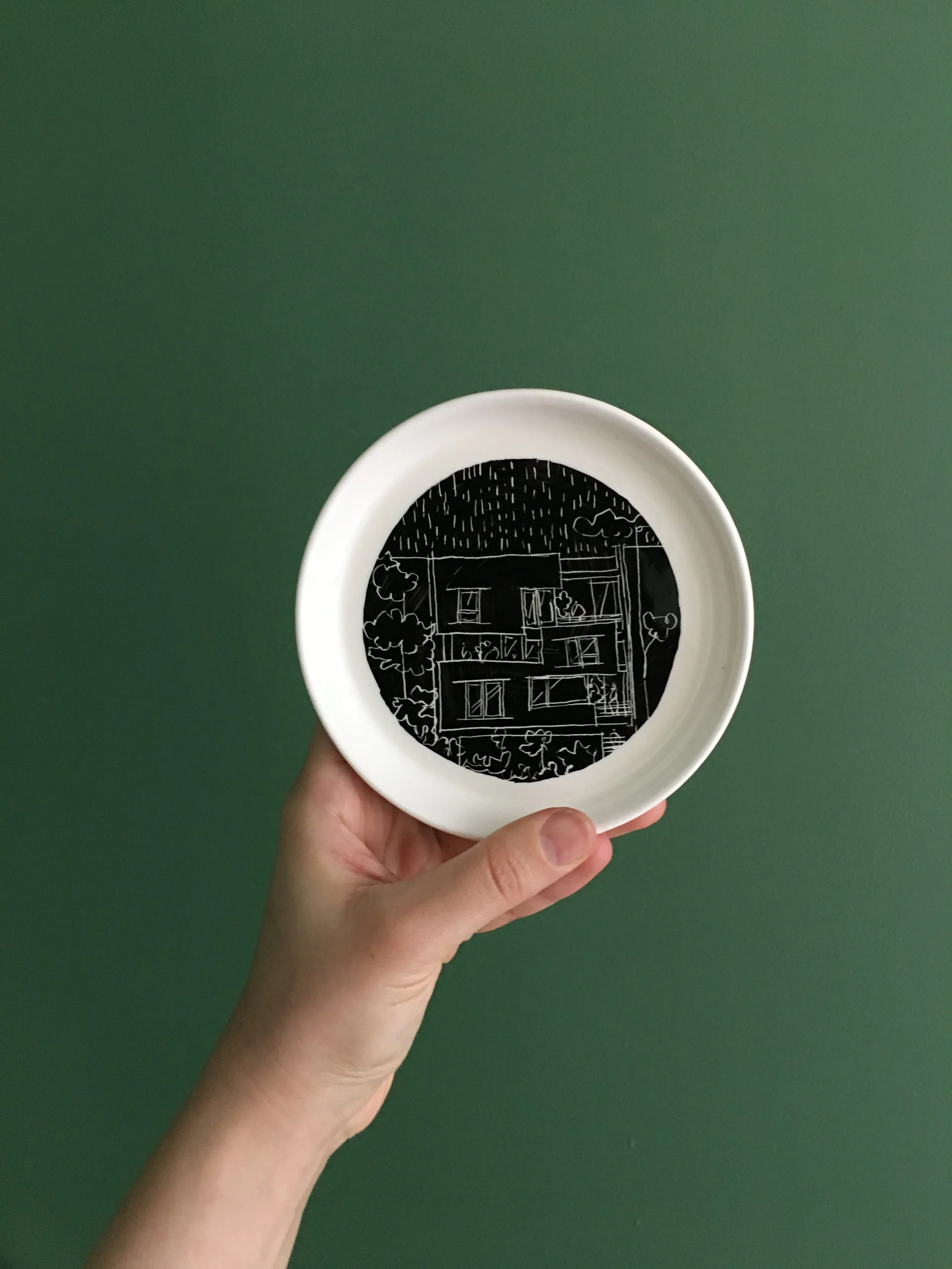 White bowl with black interior featuring a white line drawing of a house, trees, and a street scene, held in a person's hand against a green background.