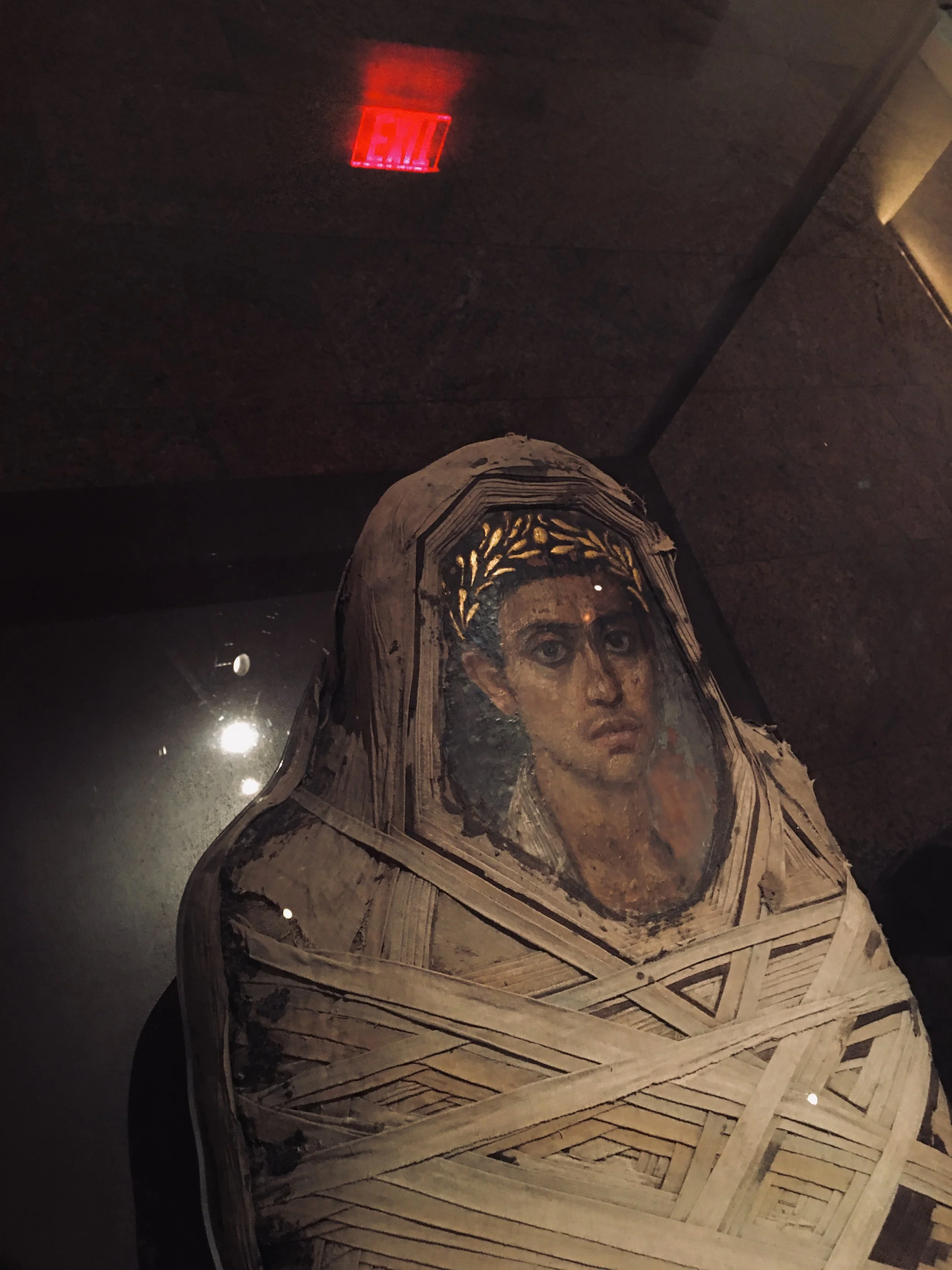 An ancient painted portrait of a woman with dark hair, wearing a golden laurel crown, wrapped in layers of cloth or bandages, onto a display case.