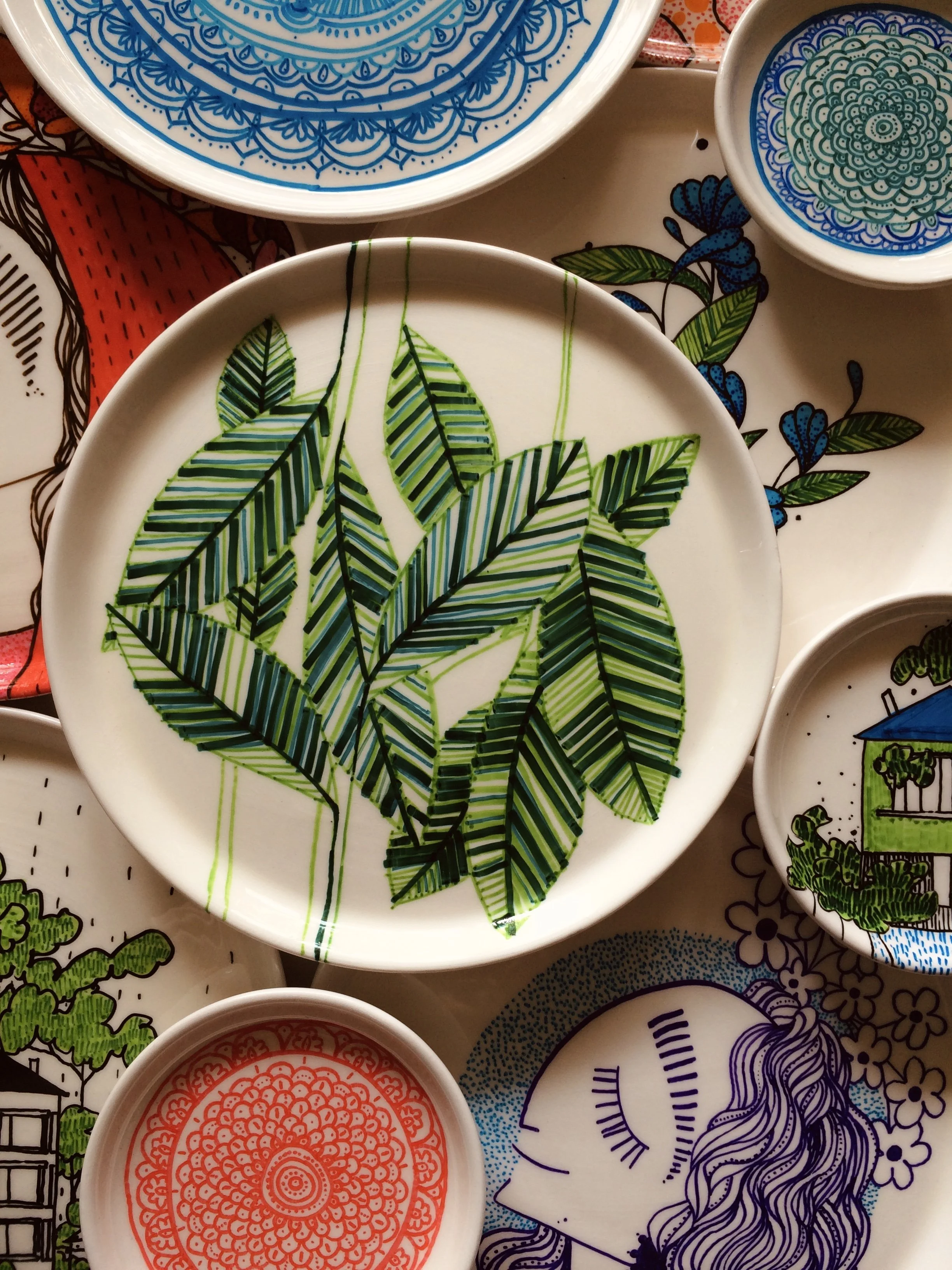 Decorative plates with abstract and stylized illustrations, including green striped leaf pattern, a girl with closed eyes and flowing hair, a house with vegetation, a detailed flower, and mandala-like designs in various colors.