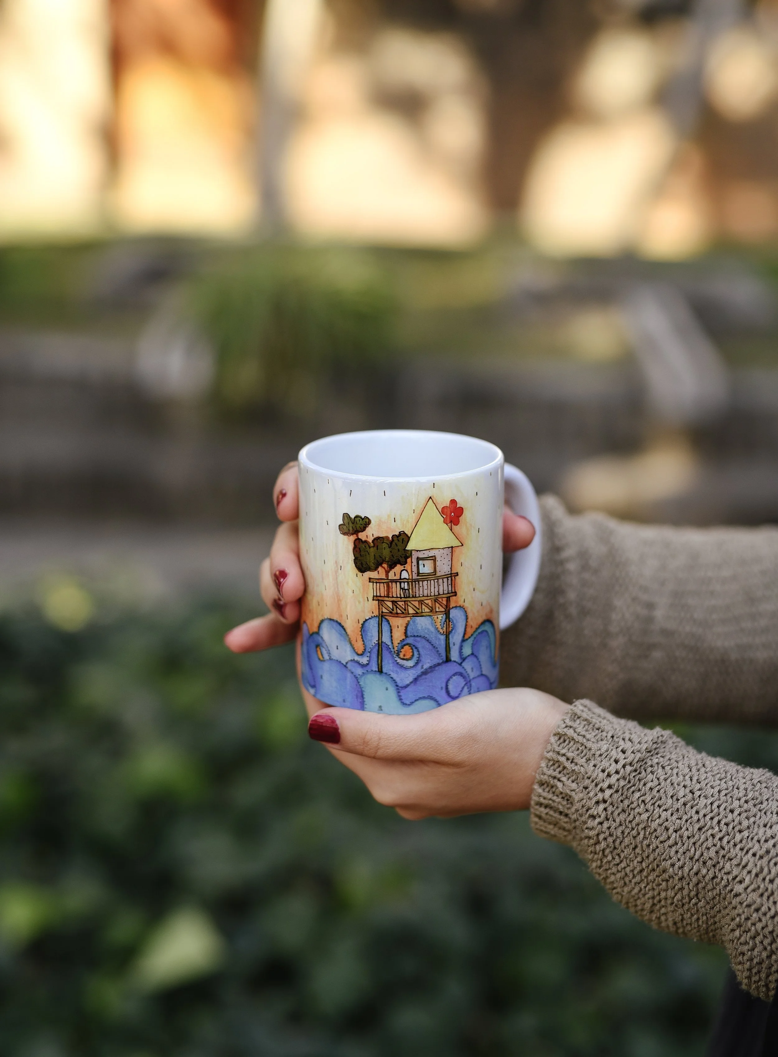 Person holding a ceramic mug with a painted scene of a house on stilts over blue water, with trees and a red flower in the background, outdoors.