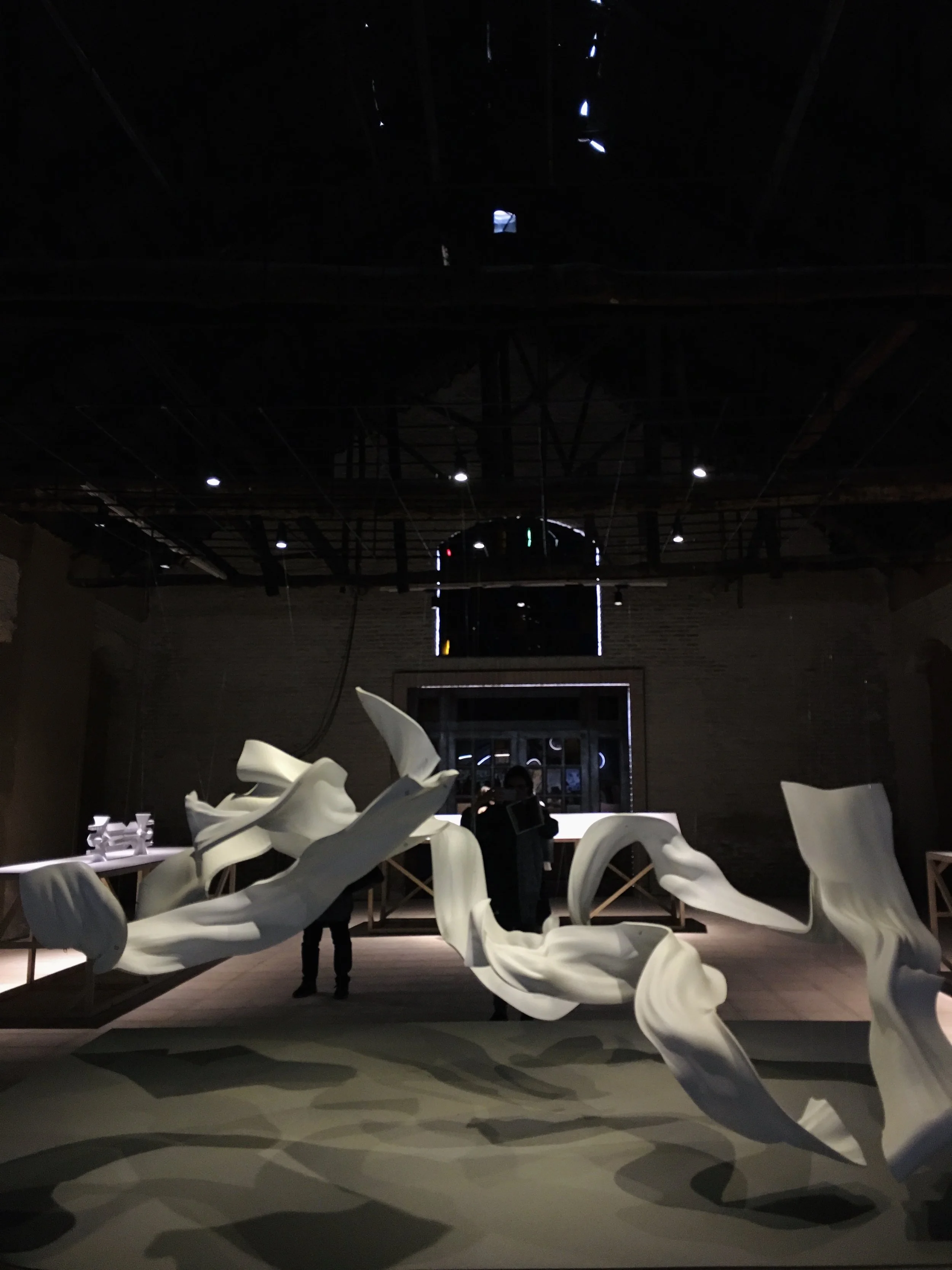 Dark room with abstract white sculptures suspended and on the floor, with a person standing in the background near a window.