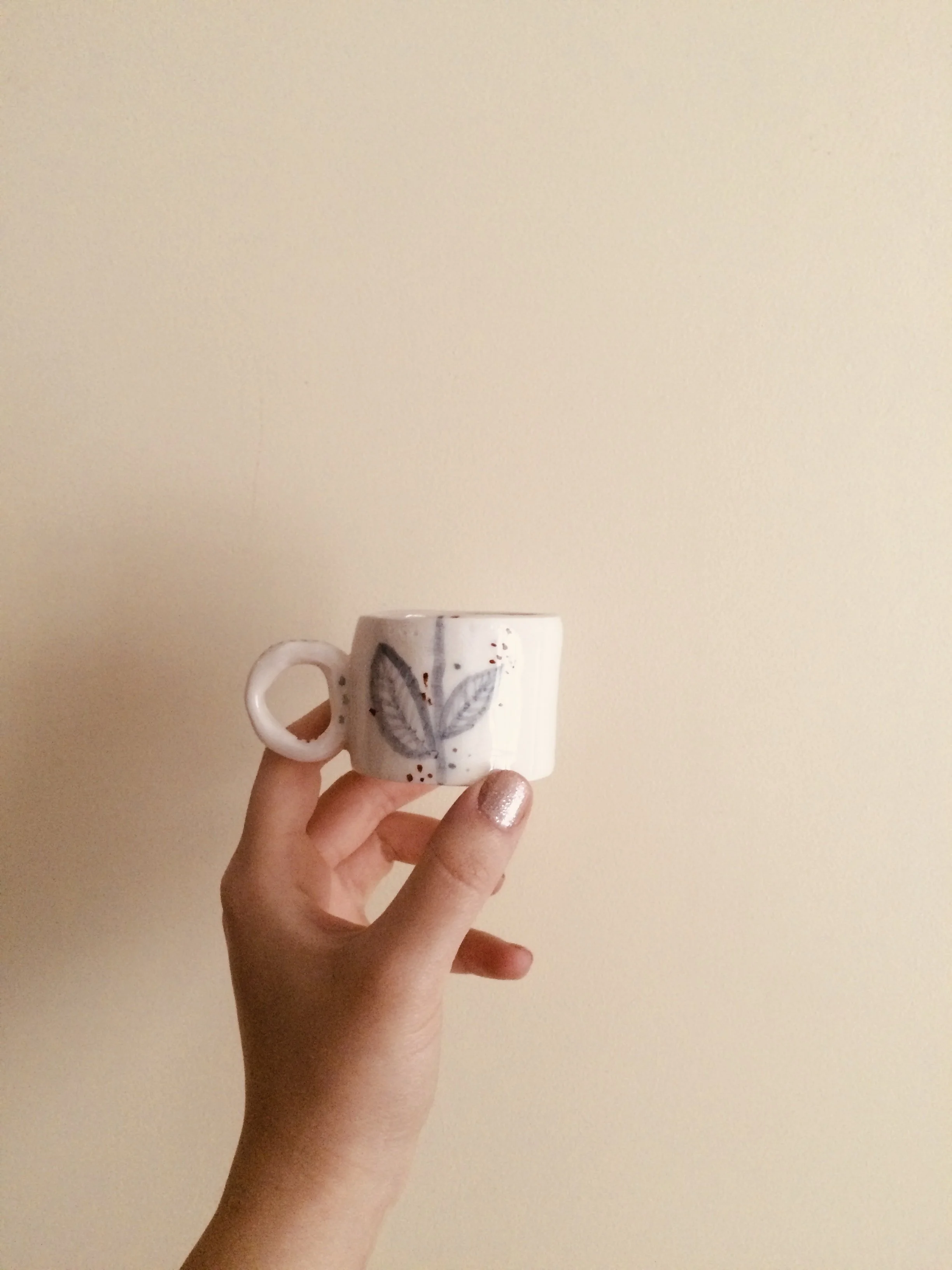 A hand holding a white ceramic mug with a painted leaf design against a plain beige wall.