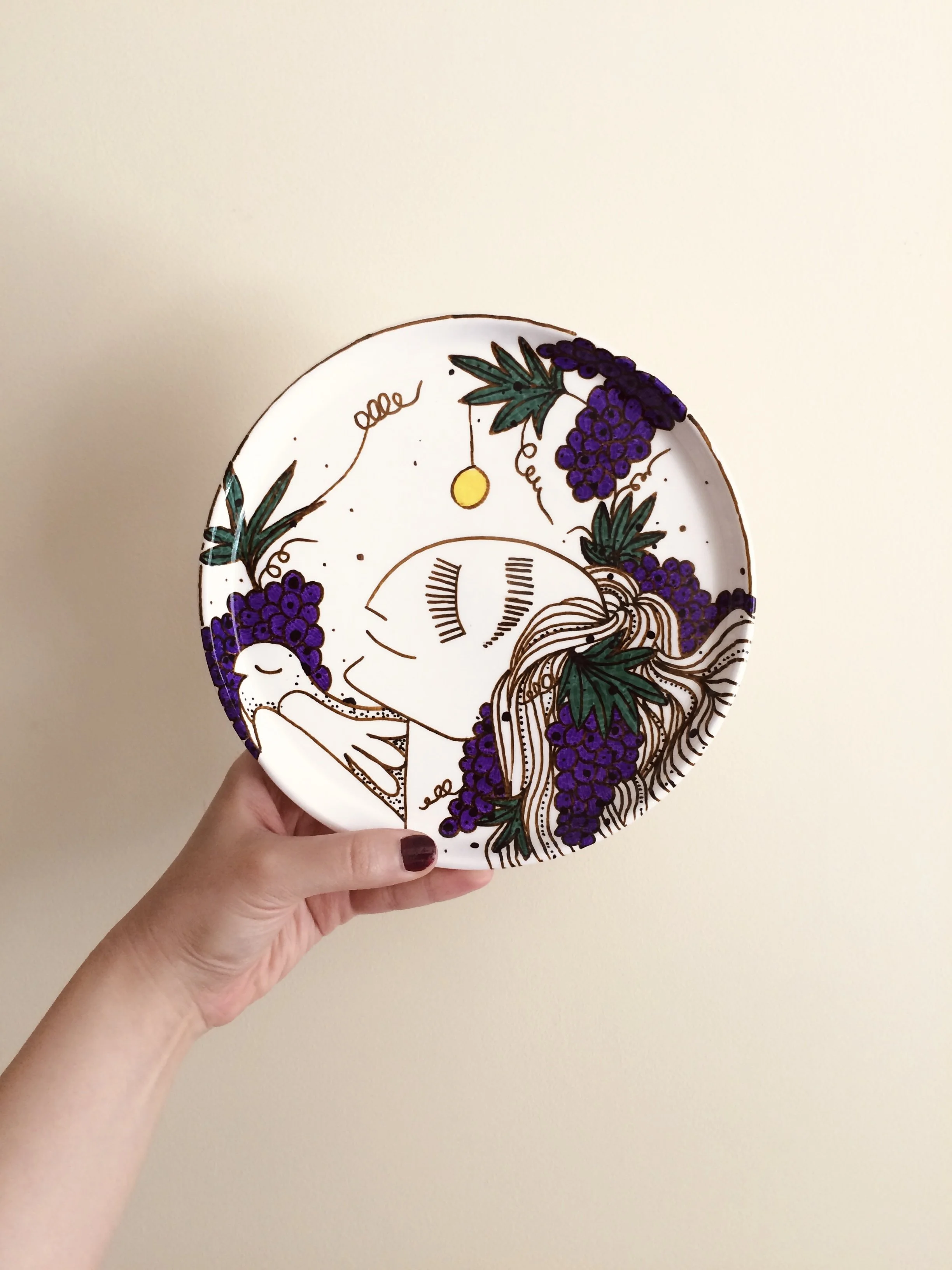 A decorative ceramic plate featuring a line drawing of a woman with closed eyes, surrounded by bunches of purple grapes and green leaves, with abstract details and a background of small black dots.