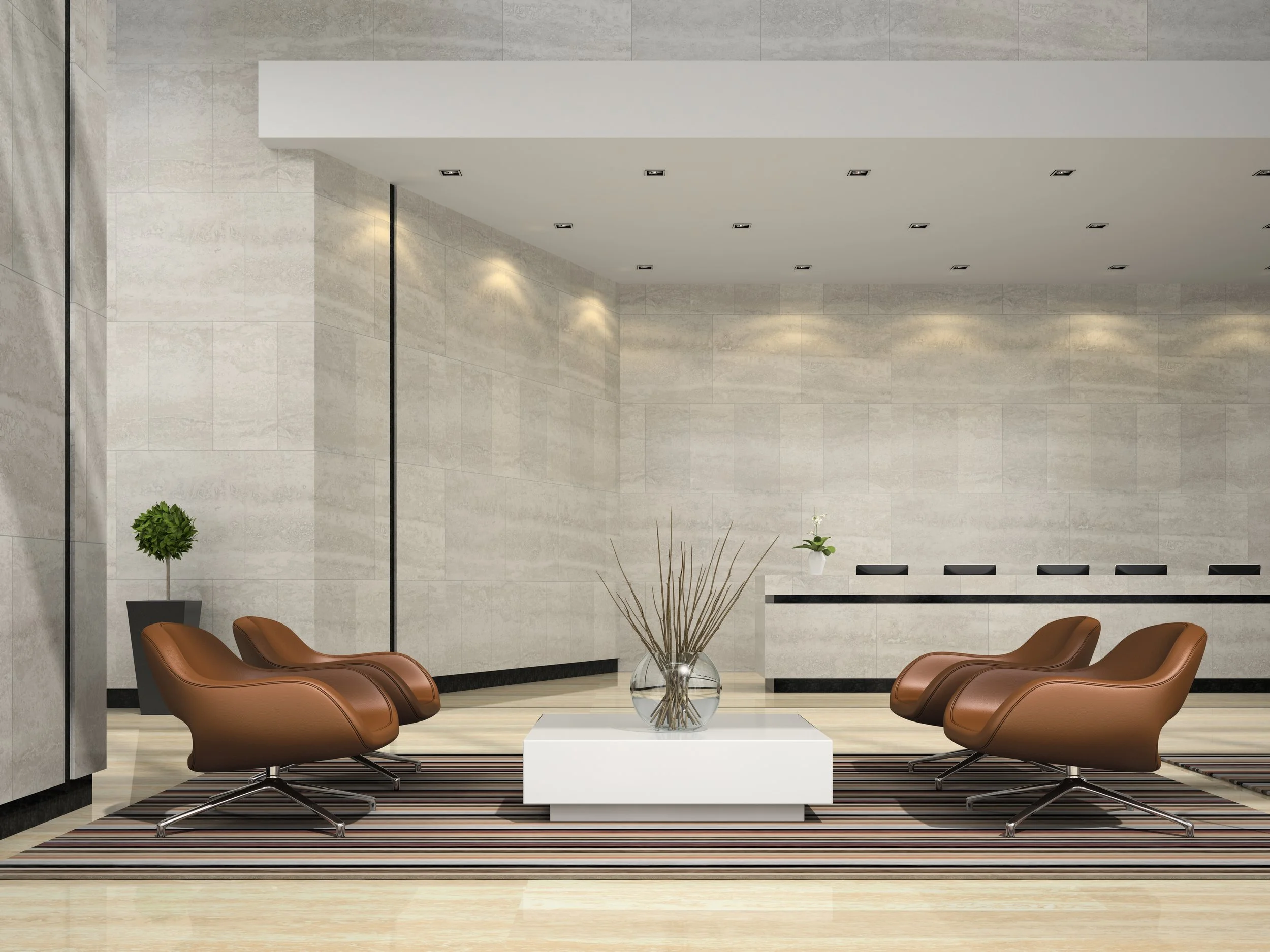 Modern hotel lobby with four brown leather chairs, a white coffee table with decorative sticks, a striped rug, a potted plant, and a reception desk in the background.