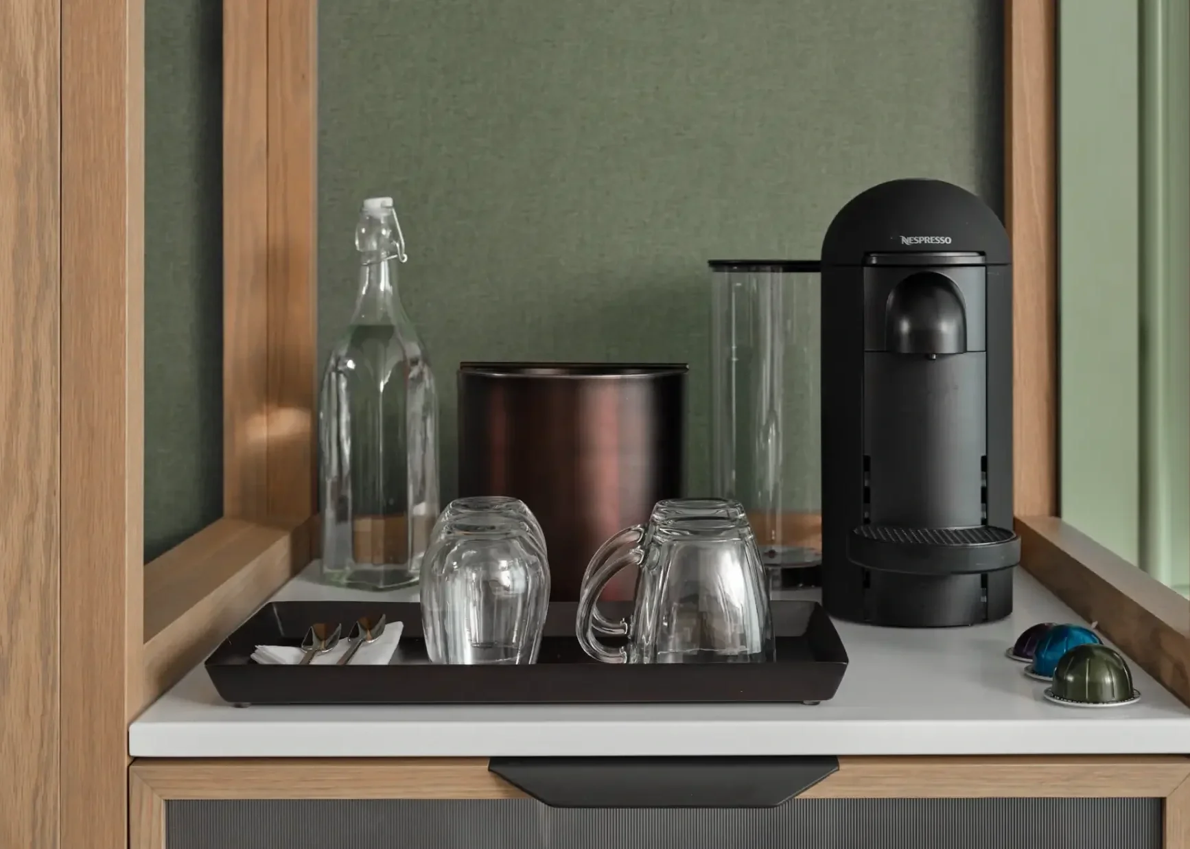 Machine à café Nespresso avec tasses en verre, bouteille en verre, boîte en métal, et capsules de différentes couleurs sur une étagère en bois.