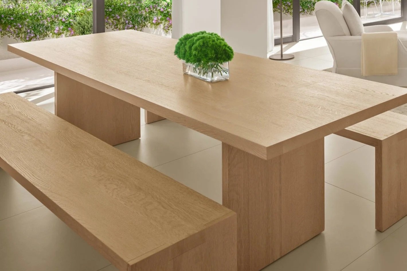 Table en bois clair avec un vase de fougères vertes, dans une salle lumineuse avec des fauteuils et une grande fenêtre.