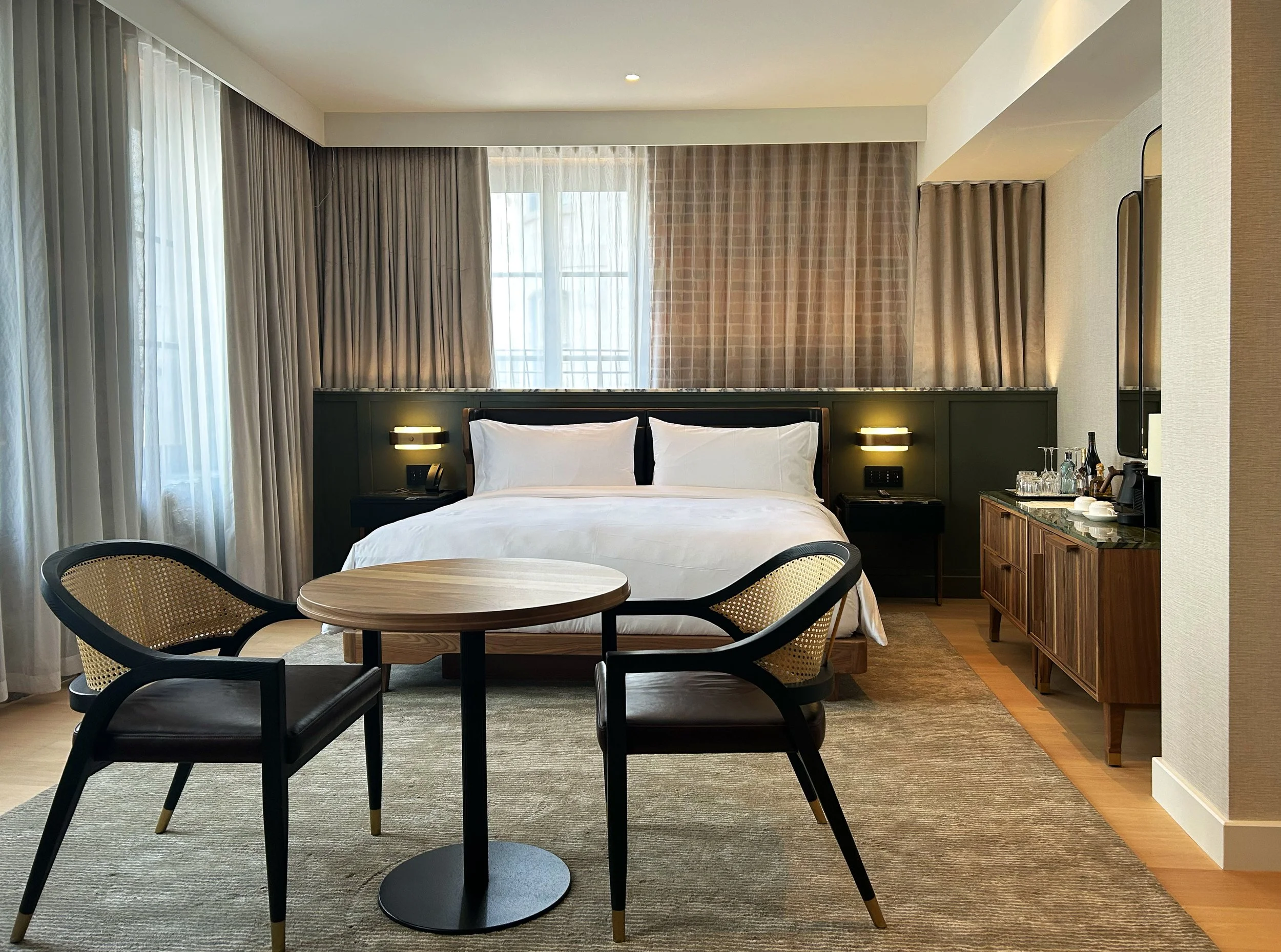 Intérieur d'une chambre d'hôtel avec un grand lit, une décoration moderne, murs en bois et canapé avec deux fauteuils, lampes sur les côtés, tableau au centre et rideaux blancs laissant entrer la lumière naturelle.