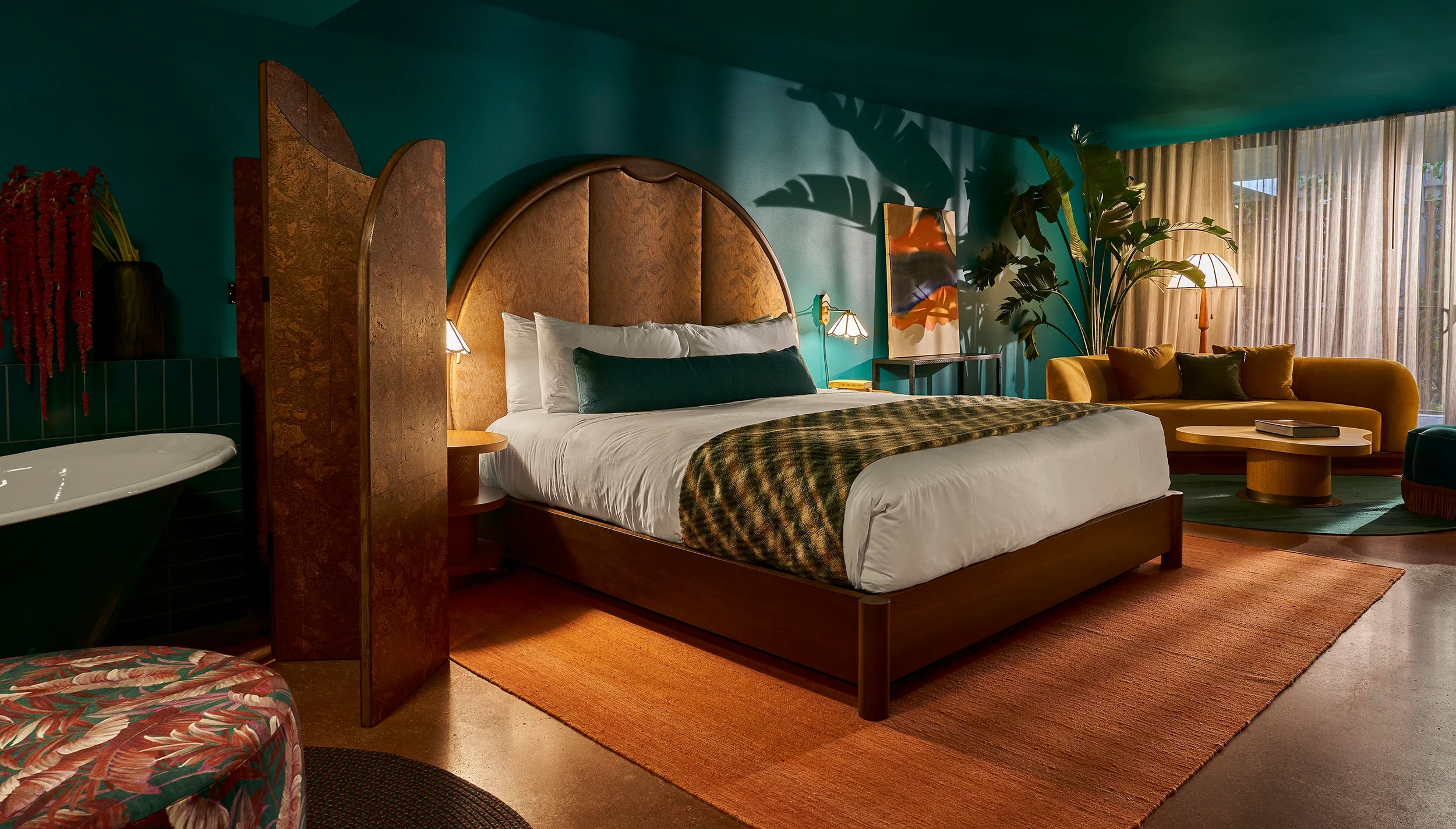 Une chambre d'hôtel élégante avec un grand lit, un canapé jaune, une table basse en bois, une grande plante, et des rideaux transparents laissant entrer la lumière naturelle.