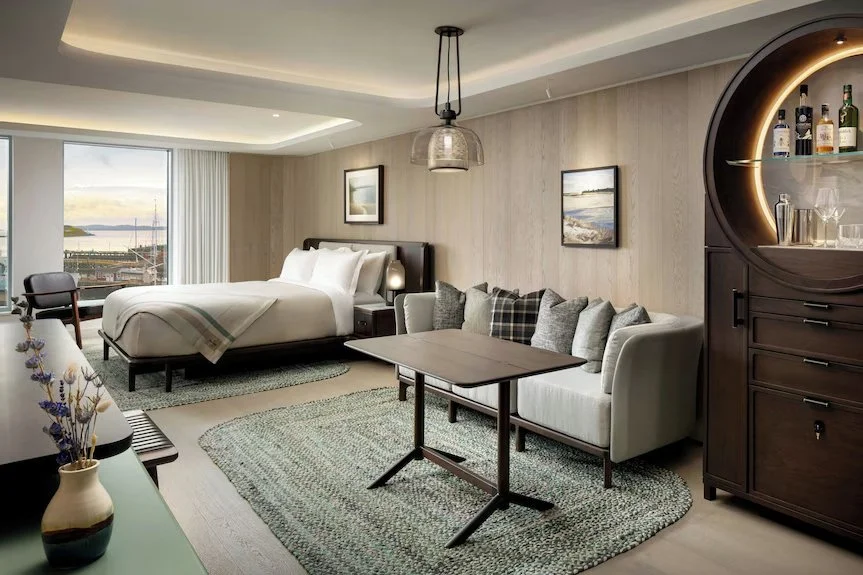 Une chambre d'hôtel moderne avec un lit double, un canapé avec des coussins, une table, un meuble avec des bouteilles d'alcool et des verres, et une grande fenêtre offrant une vue sur le port.