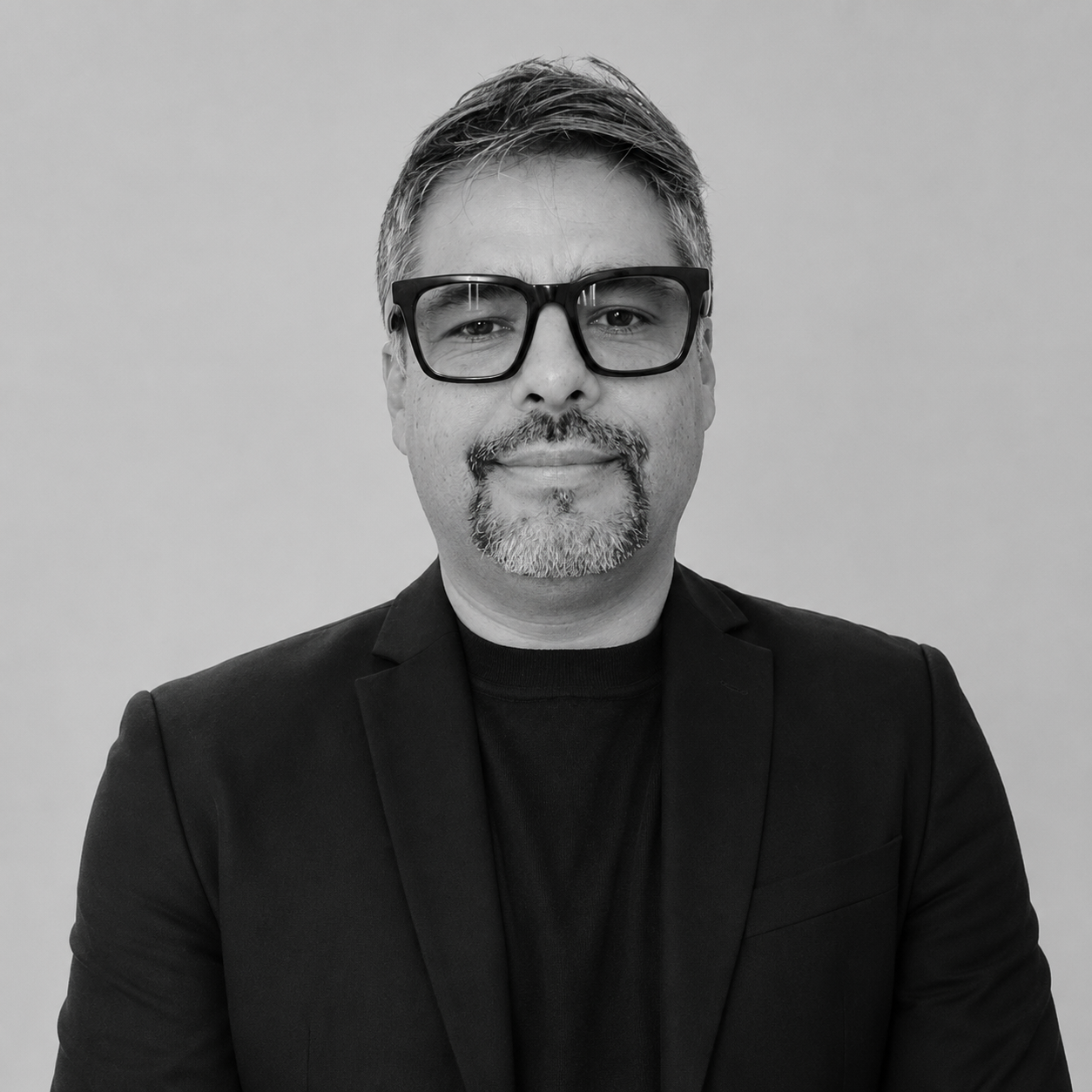 Un homme souriant avec des cheveux courts, portant un pull noir avec un logo Moncler sur la manche, photographié en noir et blanc.