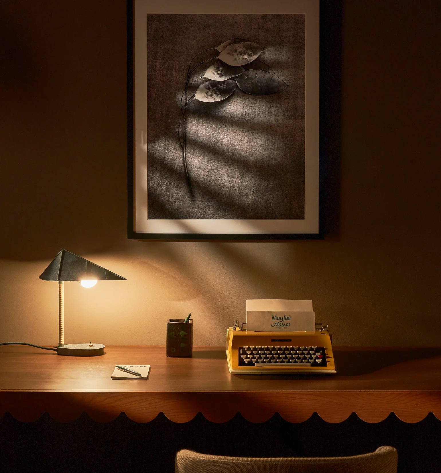 Un bureau avec une lampe, un carnet et un stylo, une machine à écrire jaune, un porte-crayons avec des stylos, et une affiche sur le mur. Il y a également un tableau noir et blanc avec des feuilles et une branche en décoration.