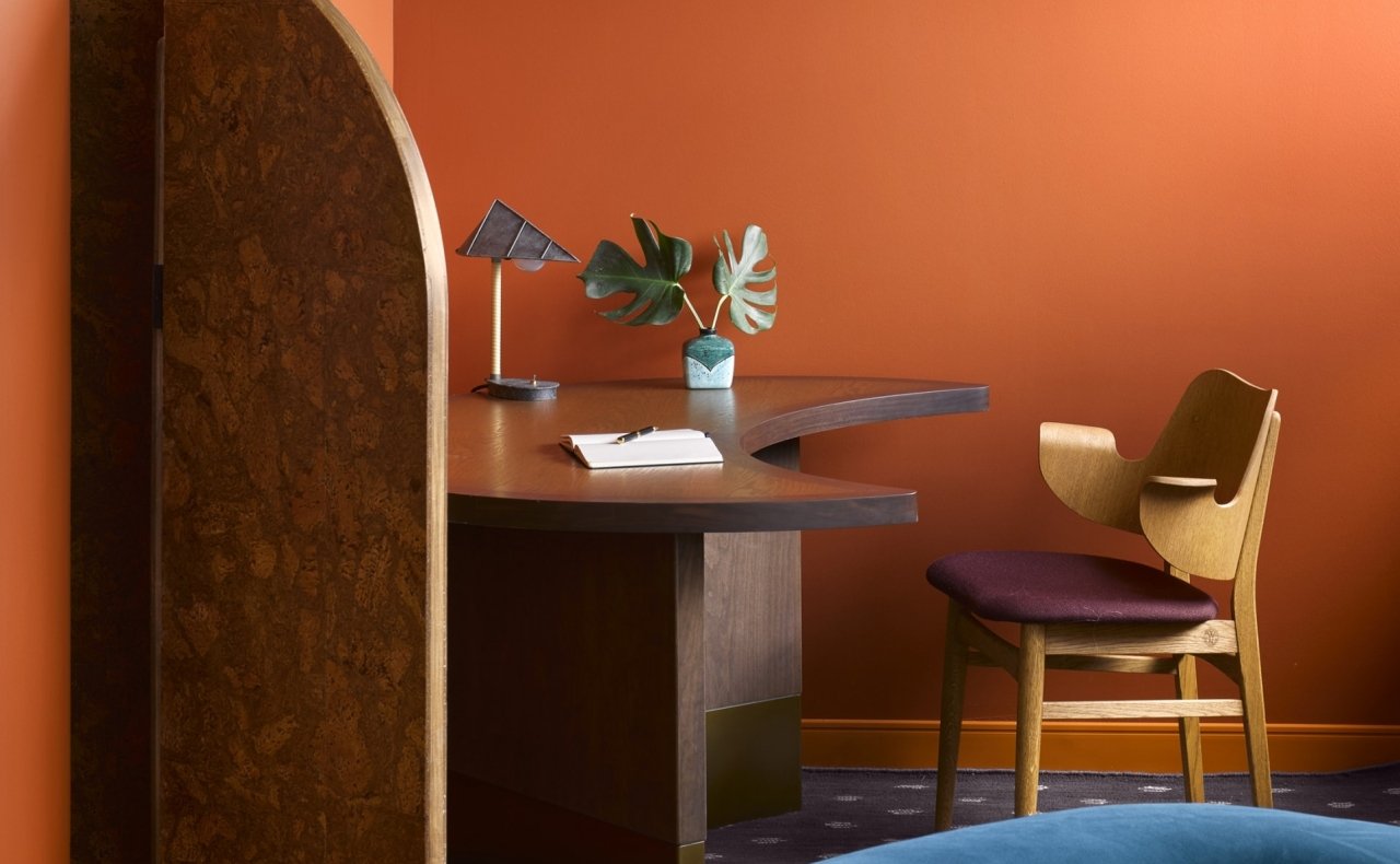Bureau en bois avec une table incurvée, une plante verte dans un vase, une lampe et un carnet, contre un mur orange, avec une chaise en bois et rembourrée de tissu violet.