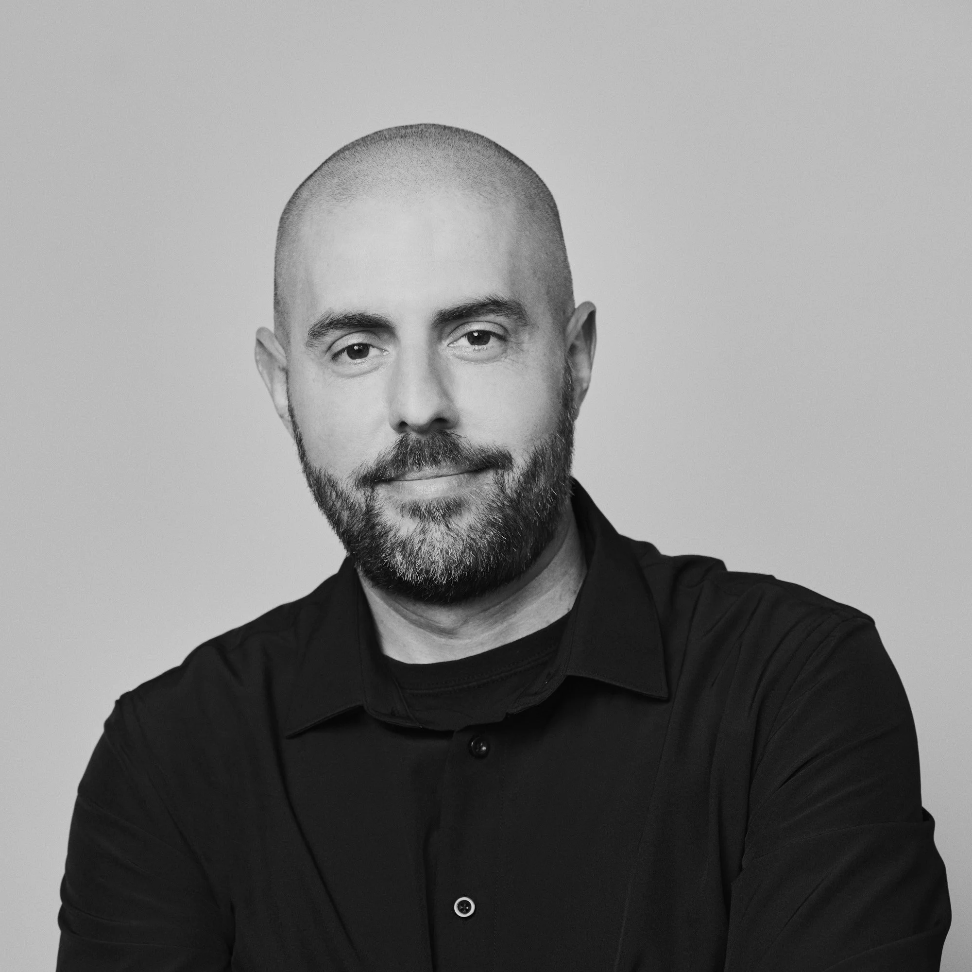 Portrait en noir et blanc d'un homme avec une barbe, portant une chemise noire, regardant vers l'objectif, avec un fond neutre.